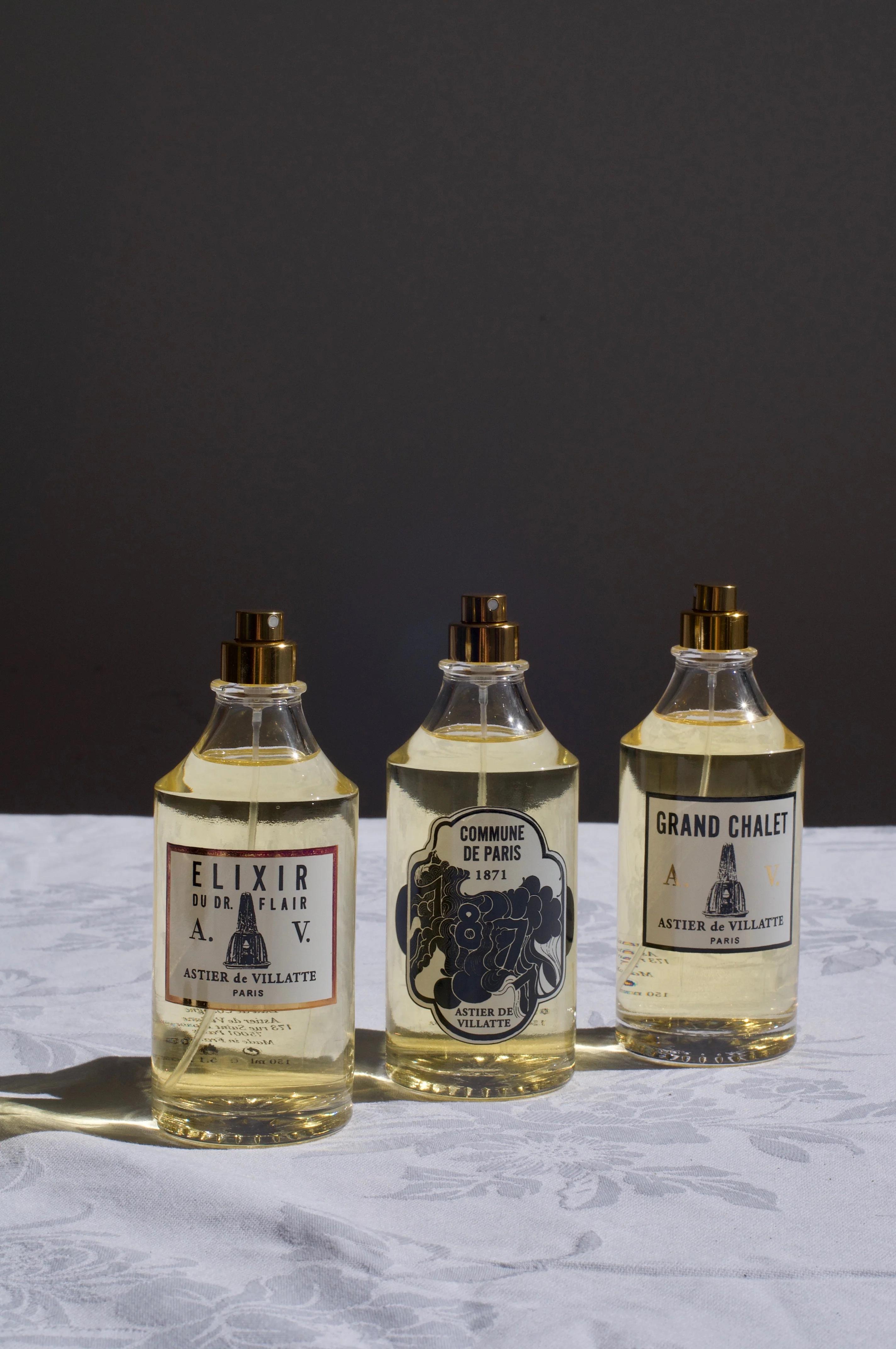 Three glass perfume bottles labeled “Elixir du Dr. Flair,” “Commune de Paris 1871,” and “Grand Chalet” on a white floral tablecloth with gold spray tops. Dark background enhances their features.