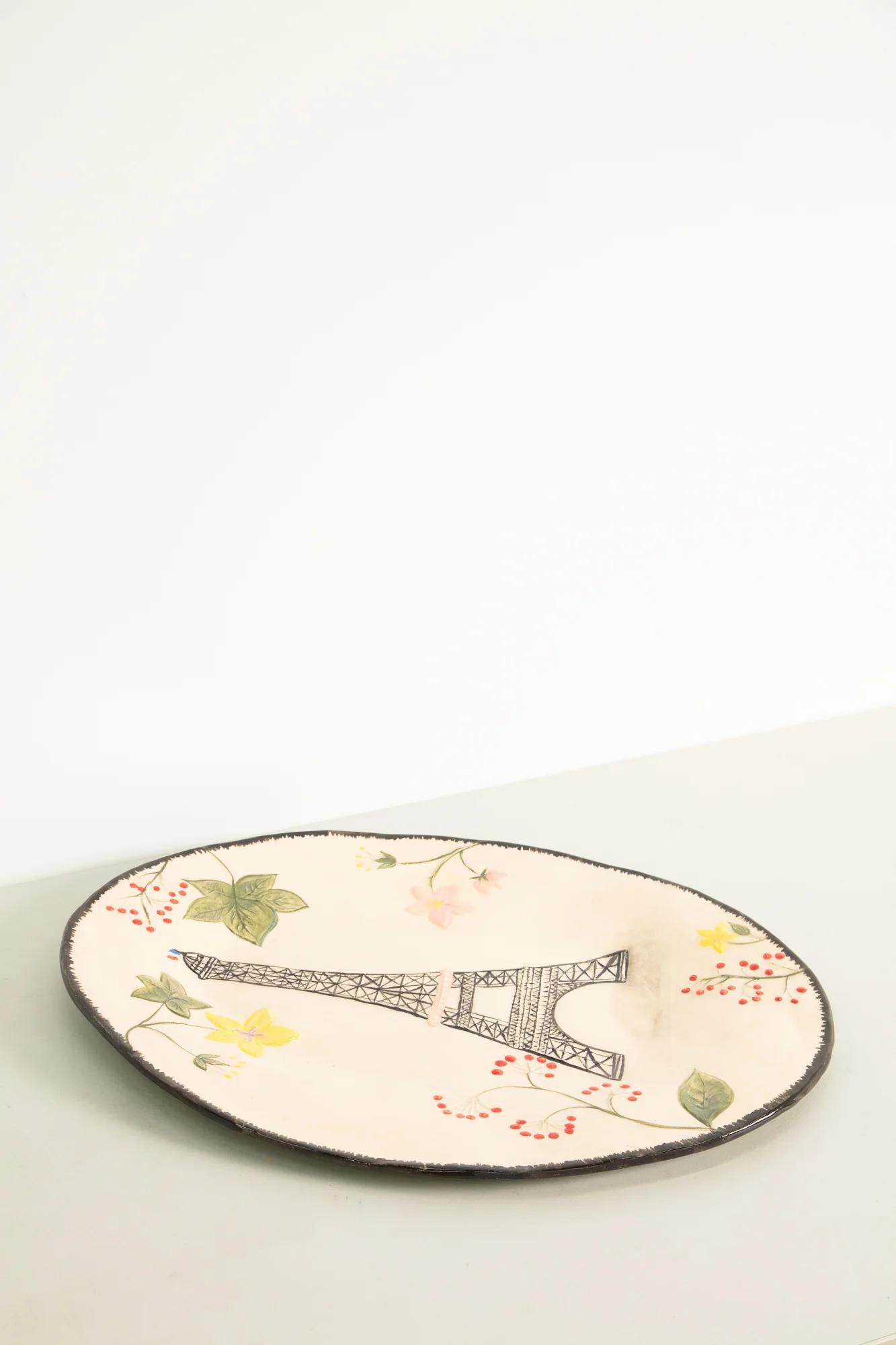 Round ceramic plate with Eiffel Tower illustration, surrounded by floral designs. White background, slightly worn black rim.