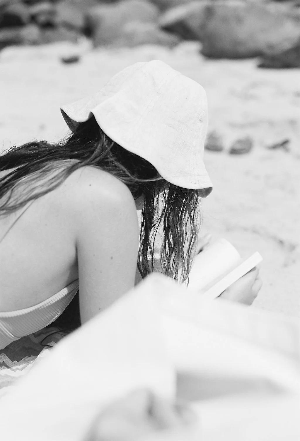 Black and white image: Long-haired person in bucket hat reading on beach. Handmade product craftsmanship subtly highlighted. Rocks in background.