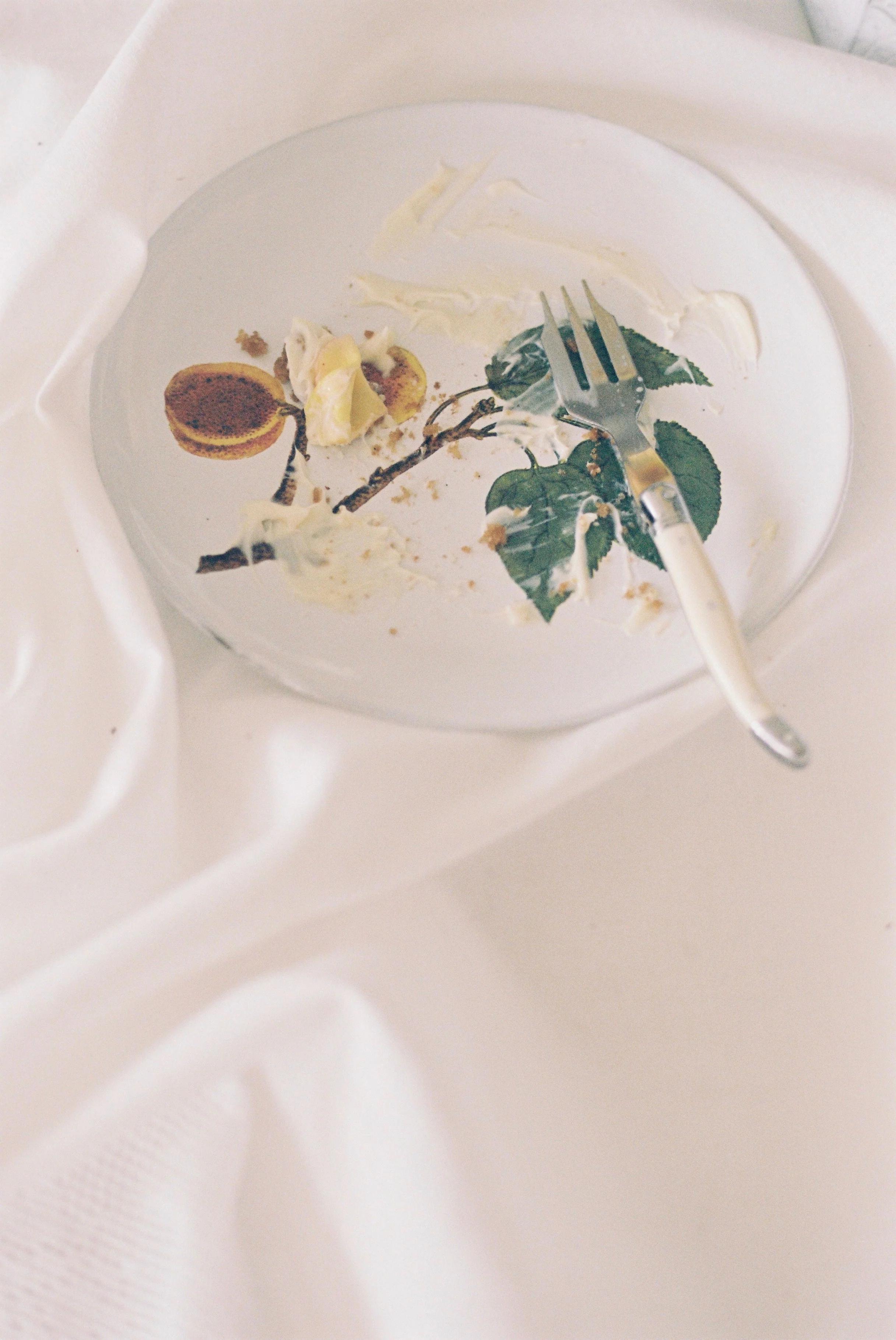 Empty floral plate with cake remnants, partially eaten tart, crumbs, and fork on crumpled tablecloth.