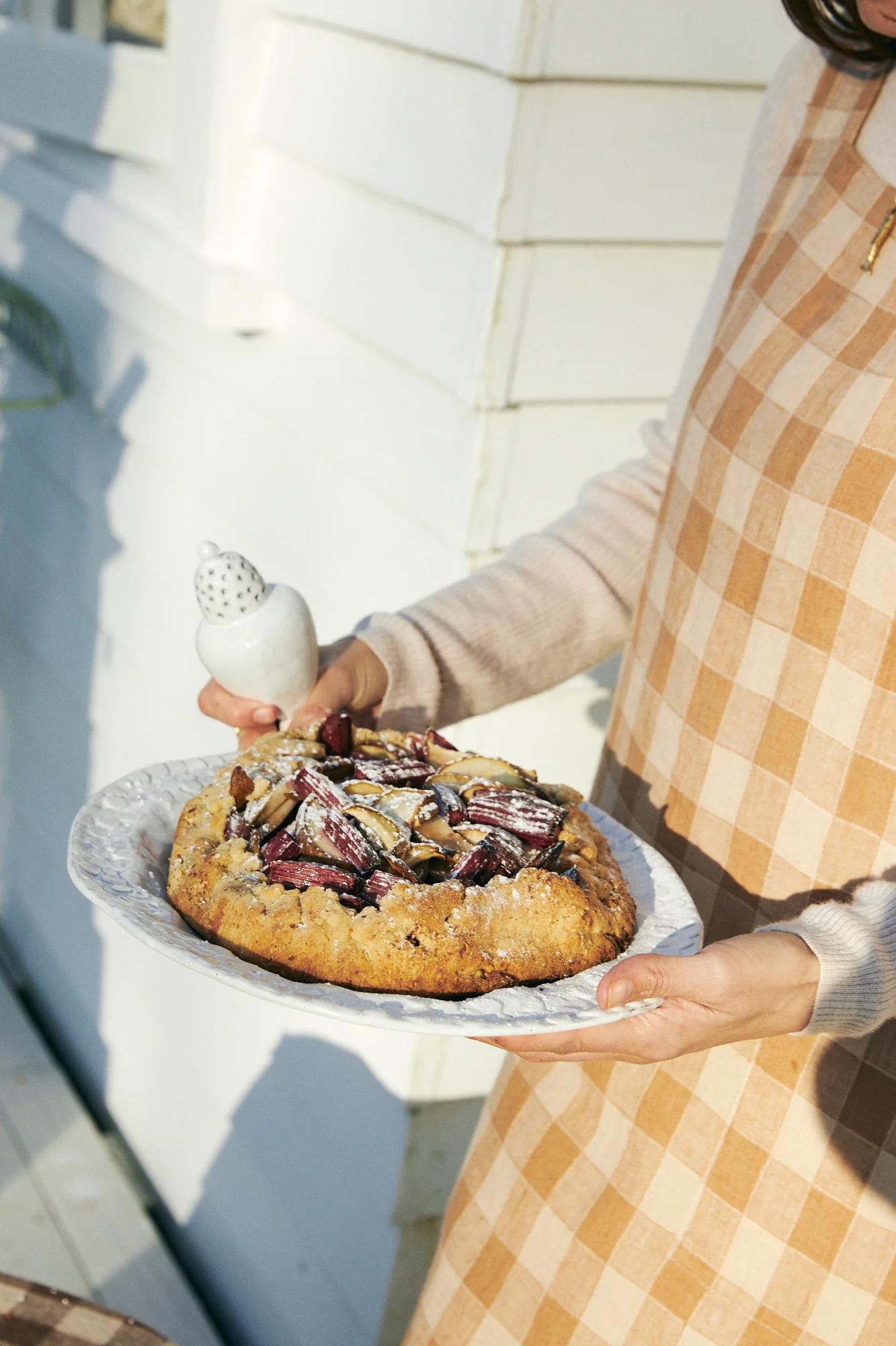 Person in checkered apron holding plate with tart and sugar shaker. White wooden wall background.