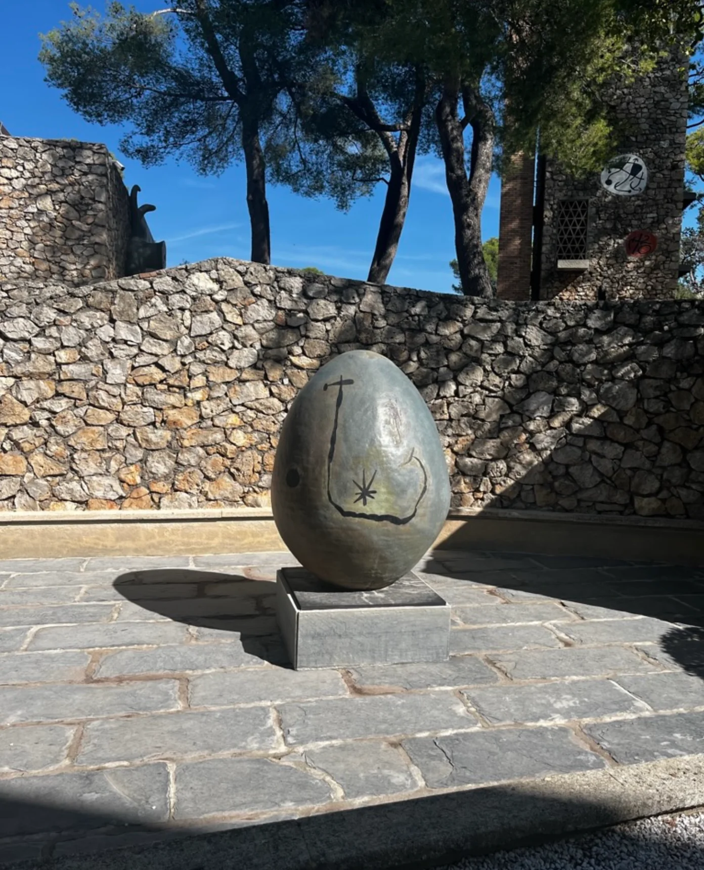 Egg-shaped stone sculpture on pedestal. Carved symbols, stone wall and trees behind. Clear blue sky.