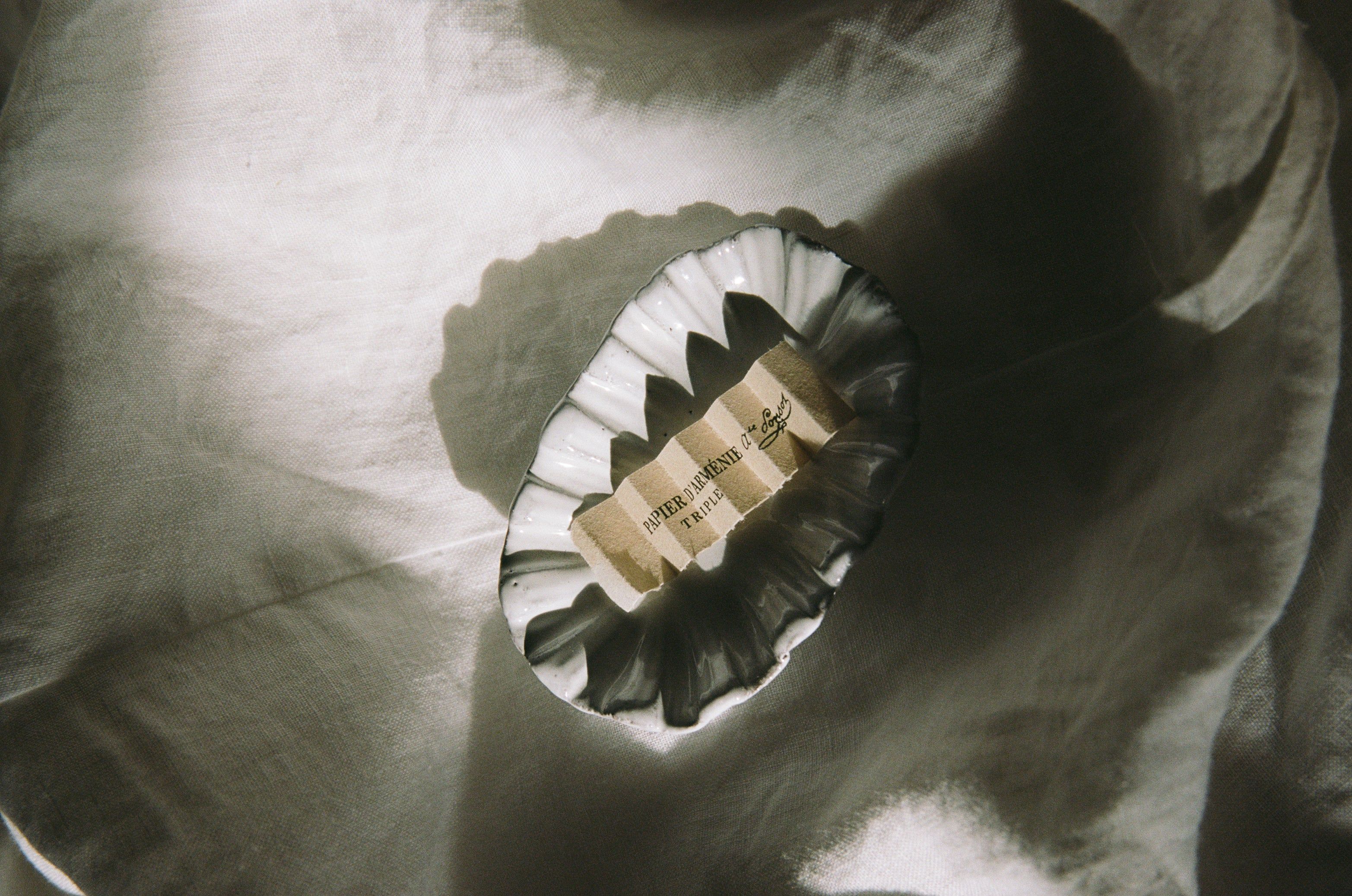 A fluted trinket dish on soft fabric holds a “Papier d’Arménie” label, partially shadowed. The dim lighting creates a dramatic ambiance.