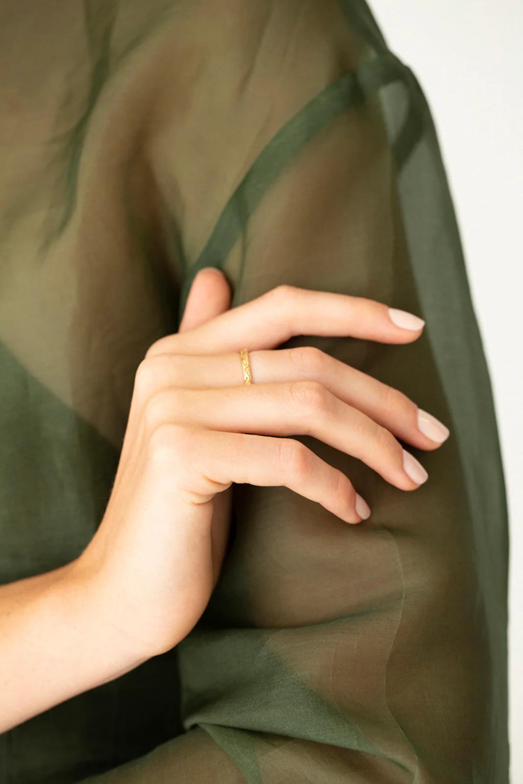 Hand with delicate Charlotte Penman gold ring. Sheer olive fabric partially covers hand. Neatly manicured nails with natural polish.