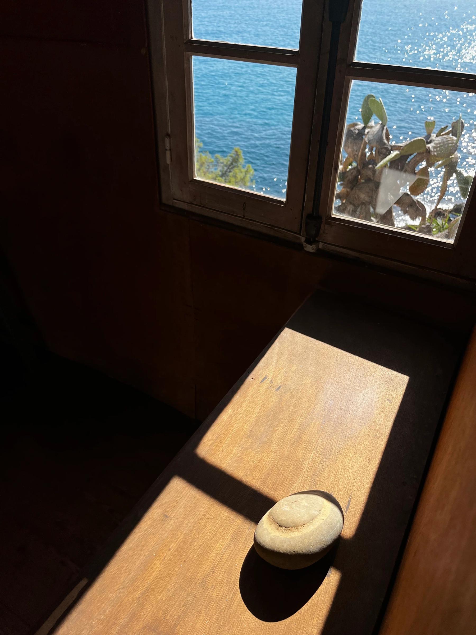 Smooth, round stone on sunlit wooden windowsill. Blue sea view, green foliage partially visible.