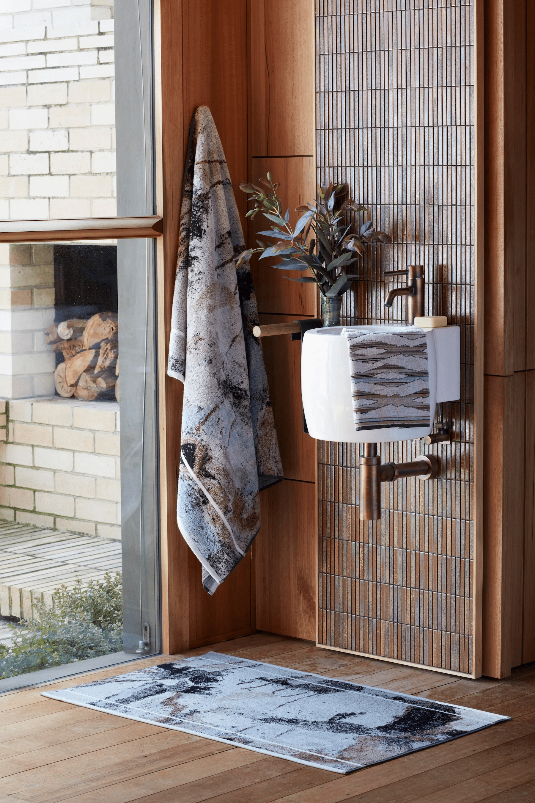 Modern bathroom with white sink, wooden walls, and tiled accents. Patterned towel and bath mat. Small plant.