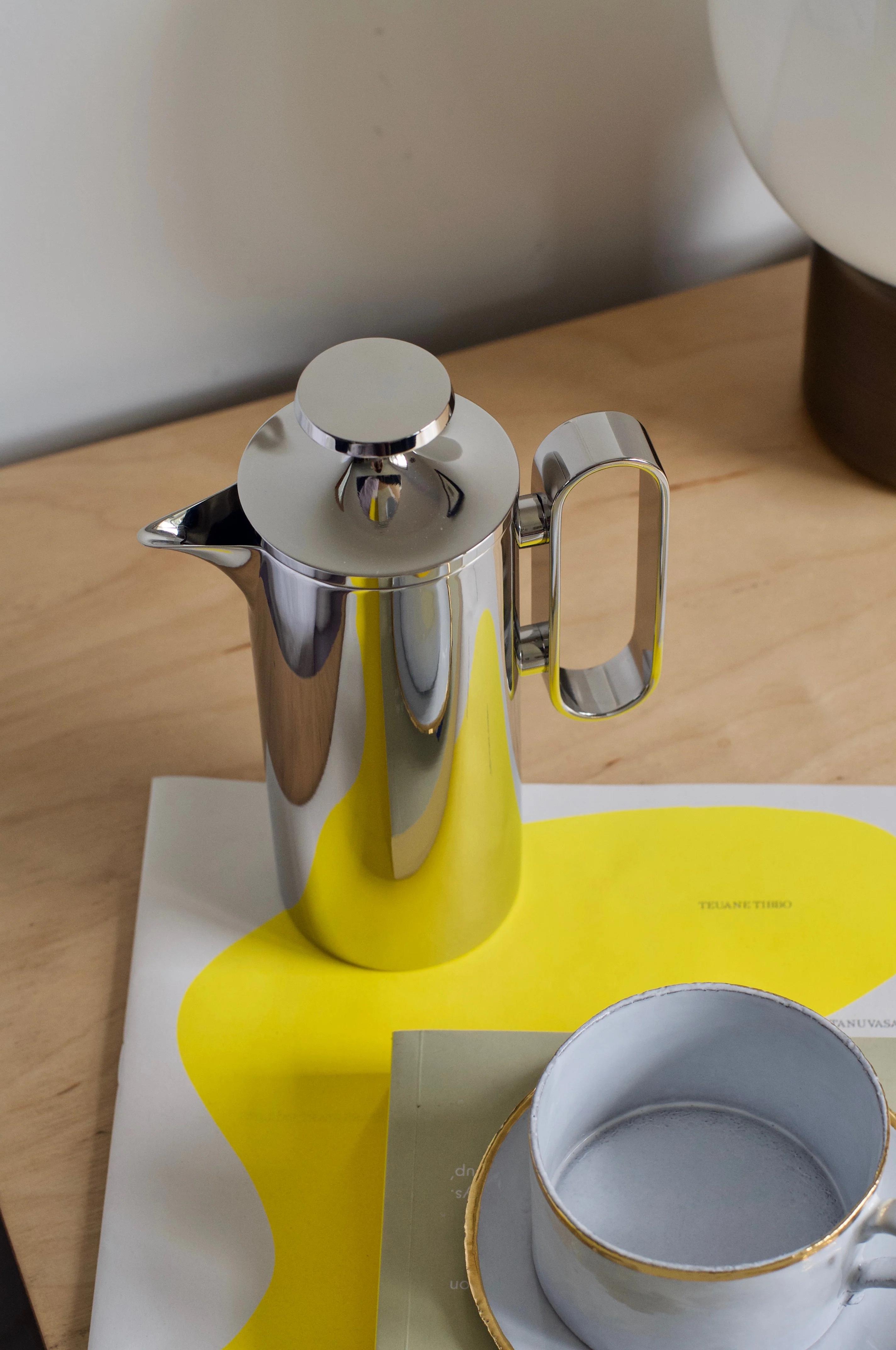 David Mellor cafetière with steel lid on wooden surface. Grey ceramic cup with gold-rimmed saucer and yellow-white magazine nearby. White lamp base in background.