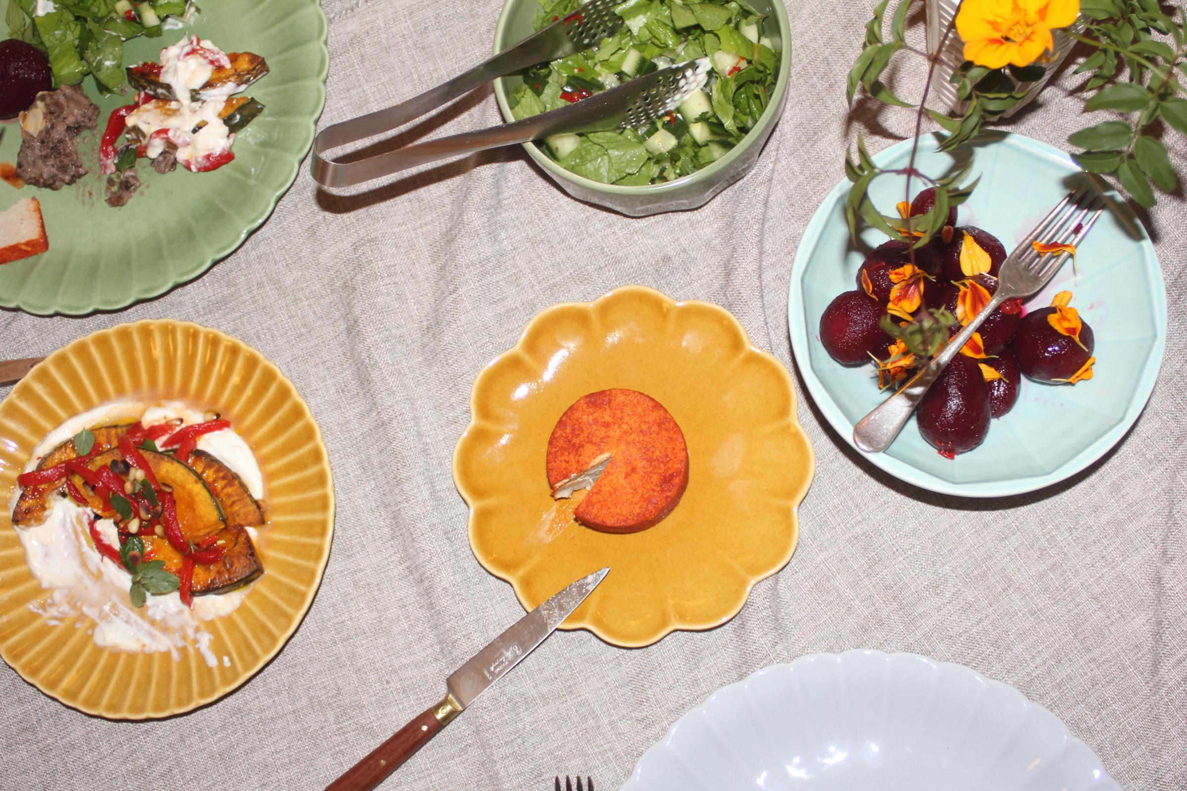Table with plated foods: orange cheese, salad, roasted veg, and flower-garnished beets. Forks and knife scattered among dishes on beige tablecloth with vibrant tableware.