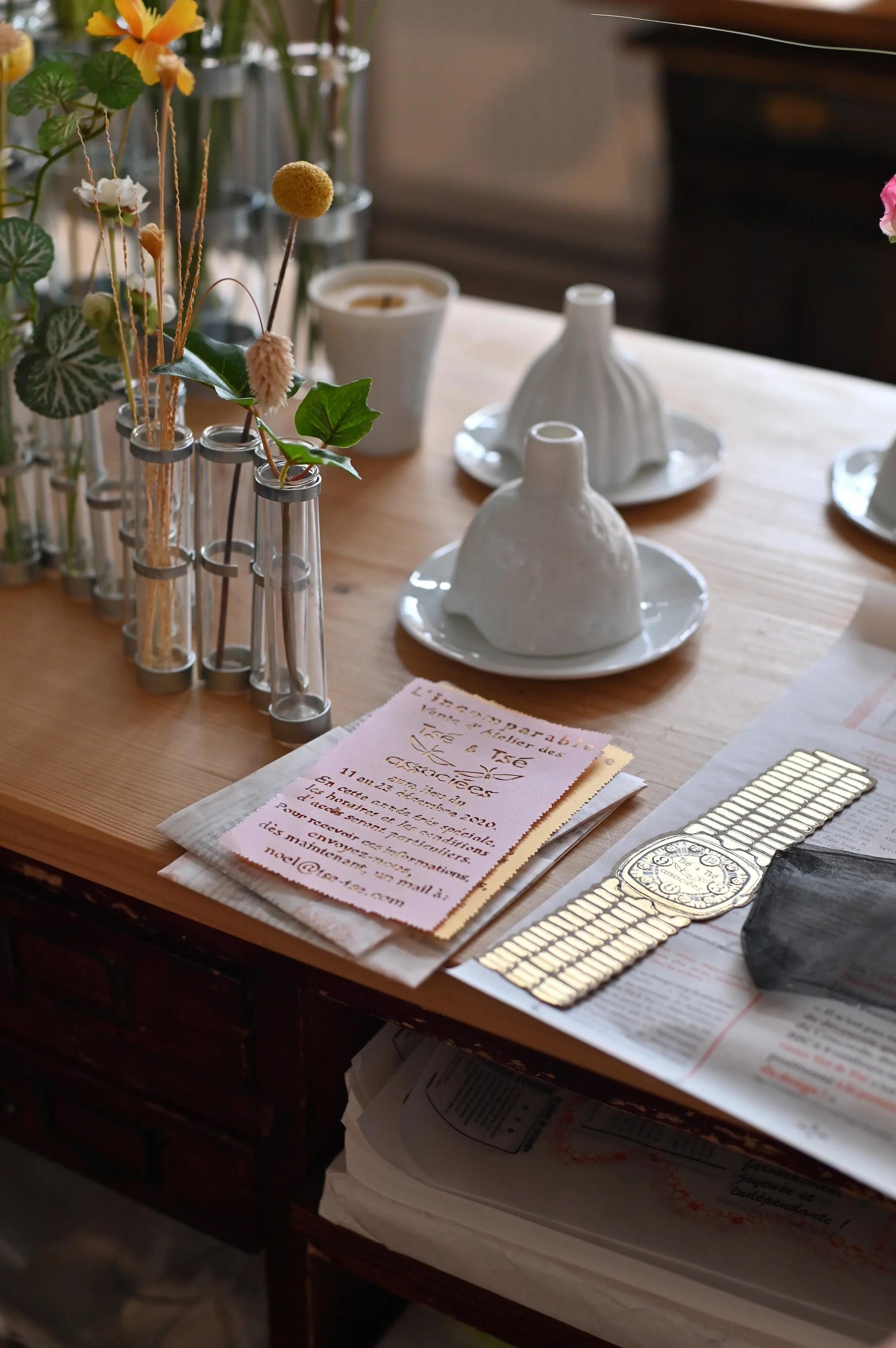 Wooden table with glass vase, ceramic cones, pink napkins, newspaper, and wristwatch. Parisian design influence evident.