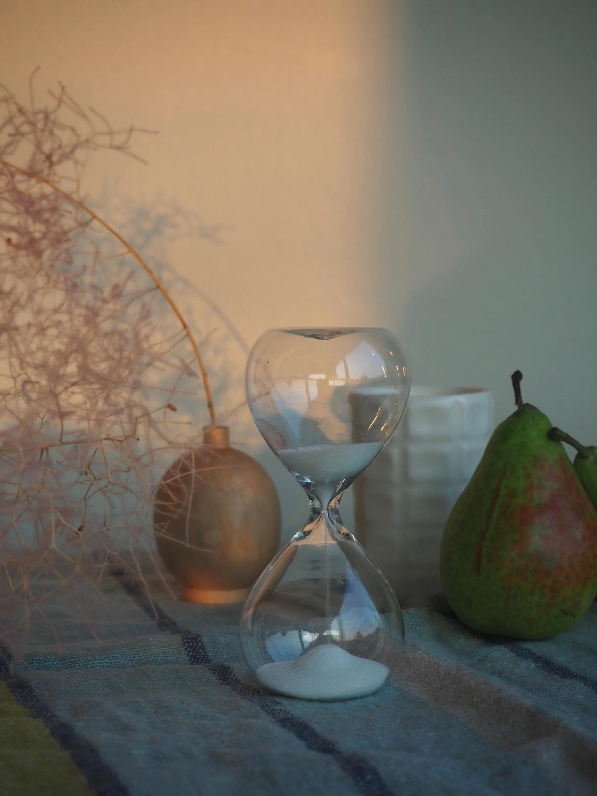 A glass hourglass, small vase, and a pear on a striped cloth, softly lit by warm dawn light, creating a peaceful morning ambiance.