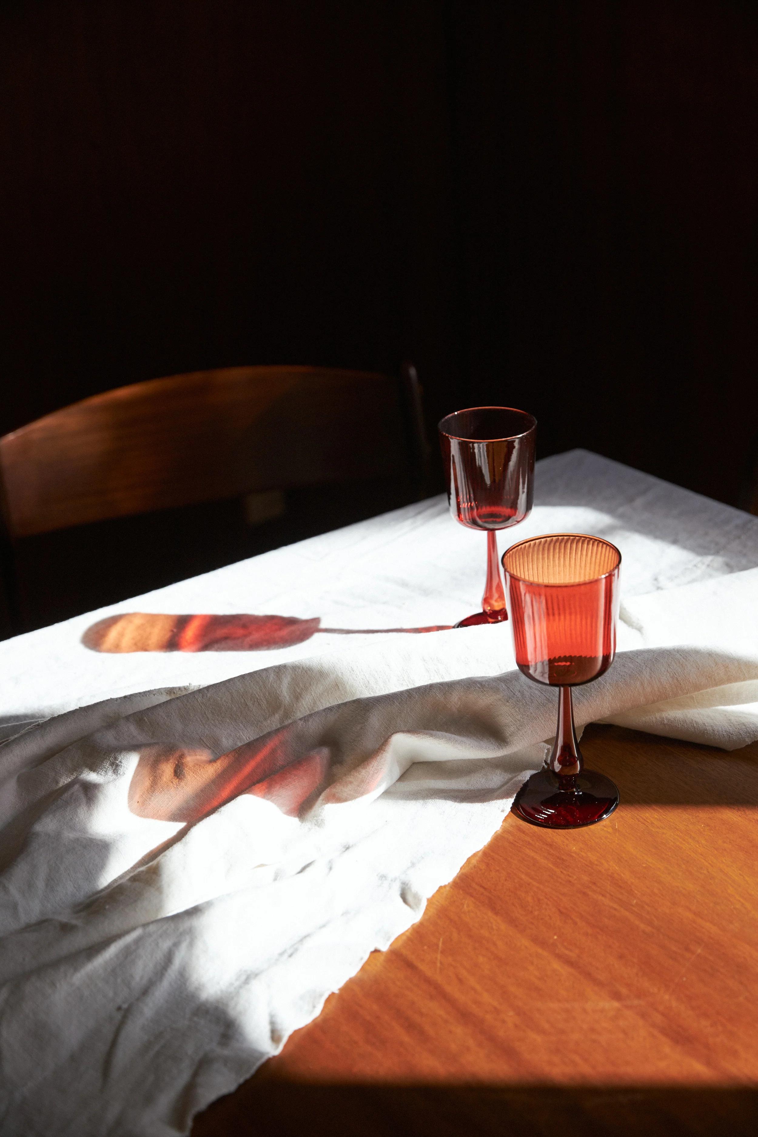 Two red wine glasses on a wooden table, partially covered by a white tablecloth. Sunlight creates shadows and reflections of the glasses on the tablecloth.