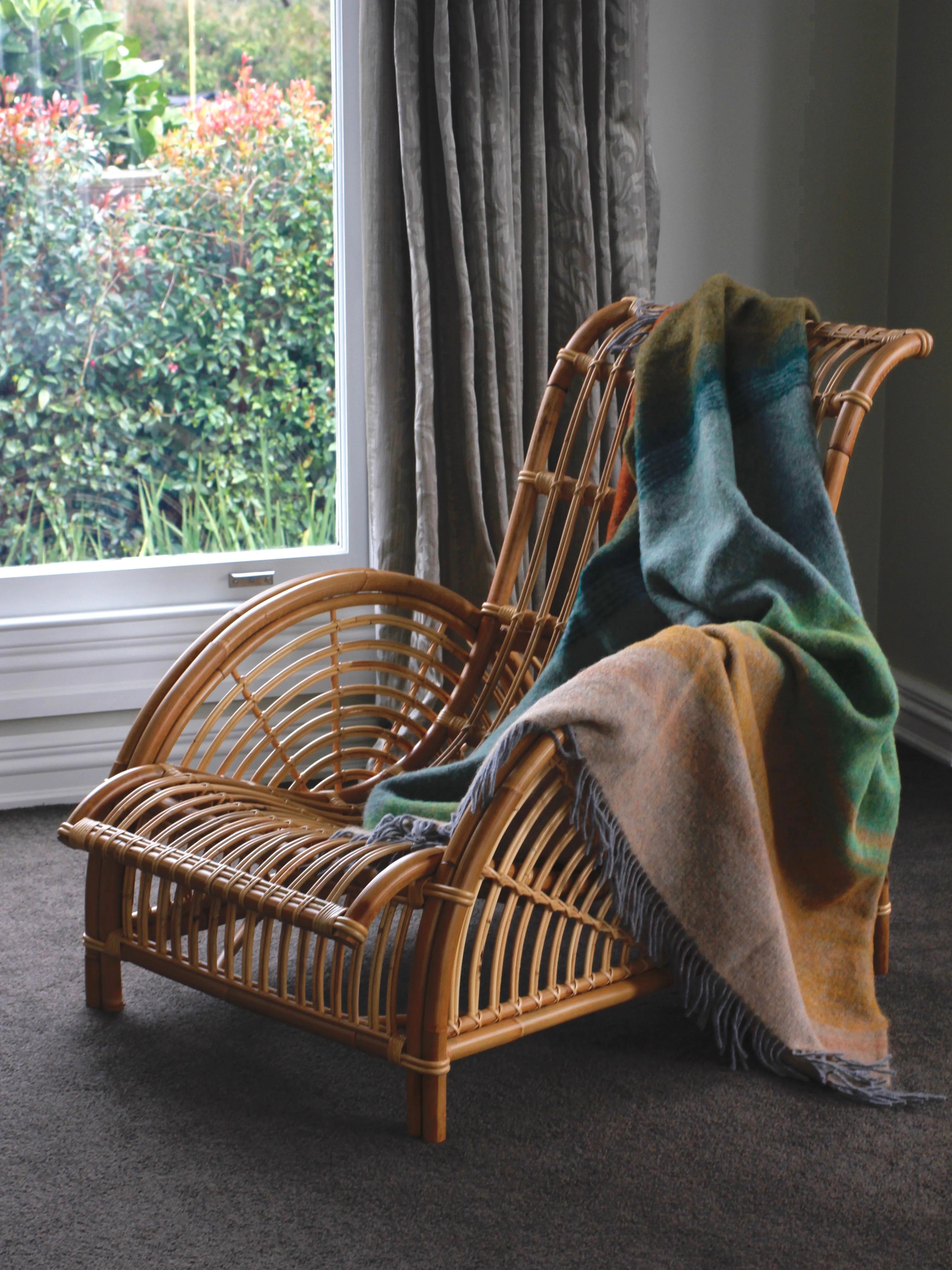 A wicker chair with a green and brown blanket, placed near a large window with grey curtains on a dark grey carpeted floor, overlooking outdoor greenery.