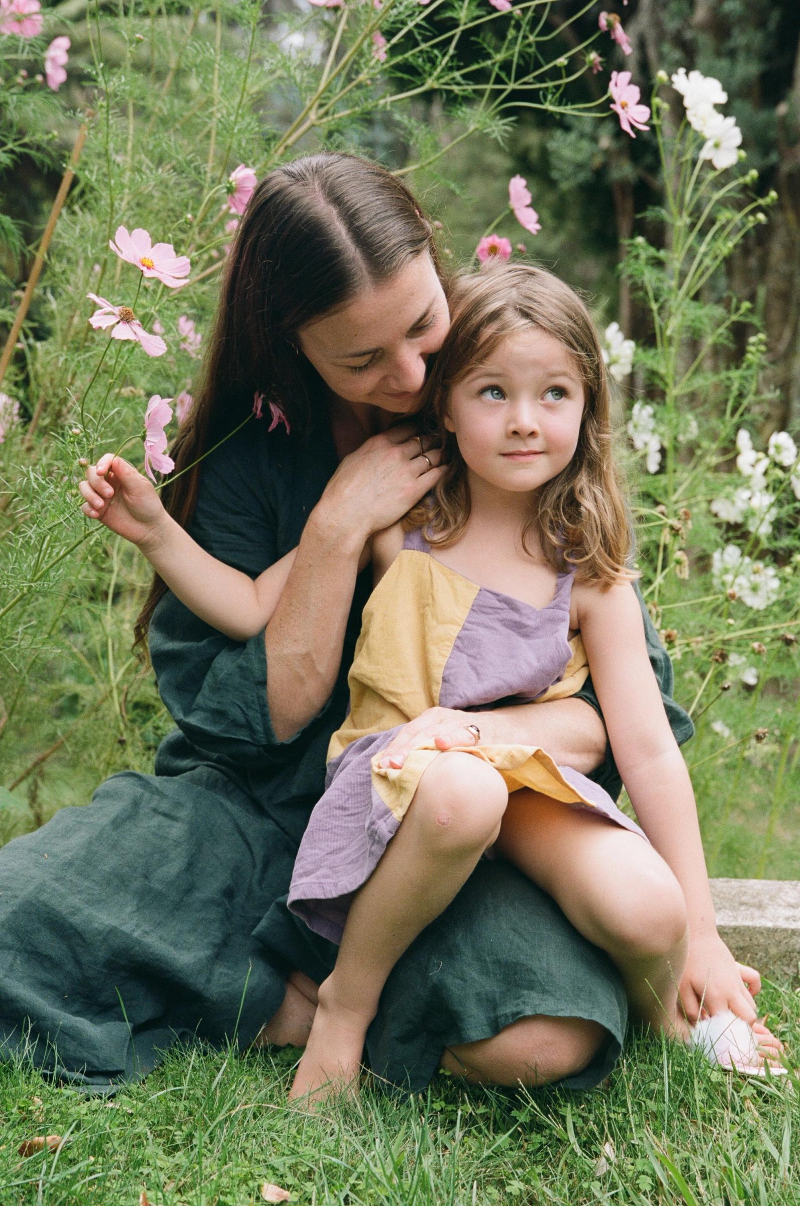 Woman embracing girl on lap in garden. Both in casual dresses. Girl holding pink flower. Green foliage background.