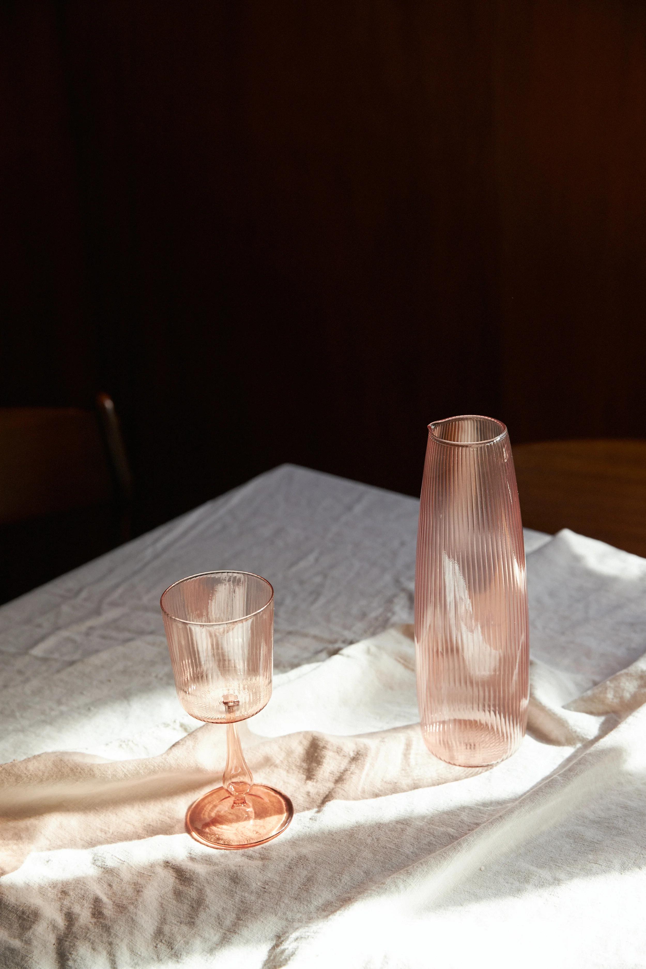 A pink goblet and ribbed carafe on a white tablecloth. Sunlight casts shadows and creates reflections of the pink glassware on the fabric.