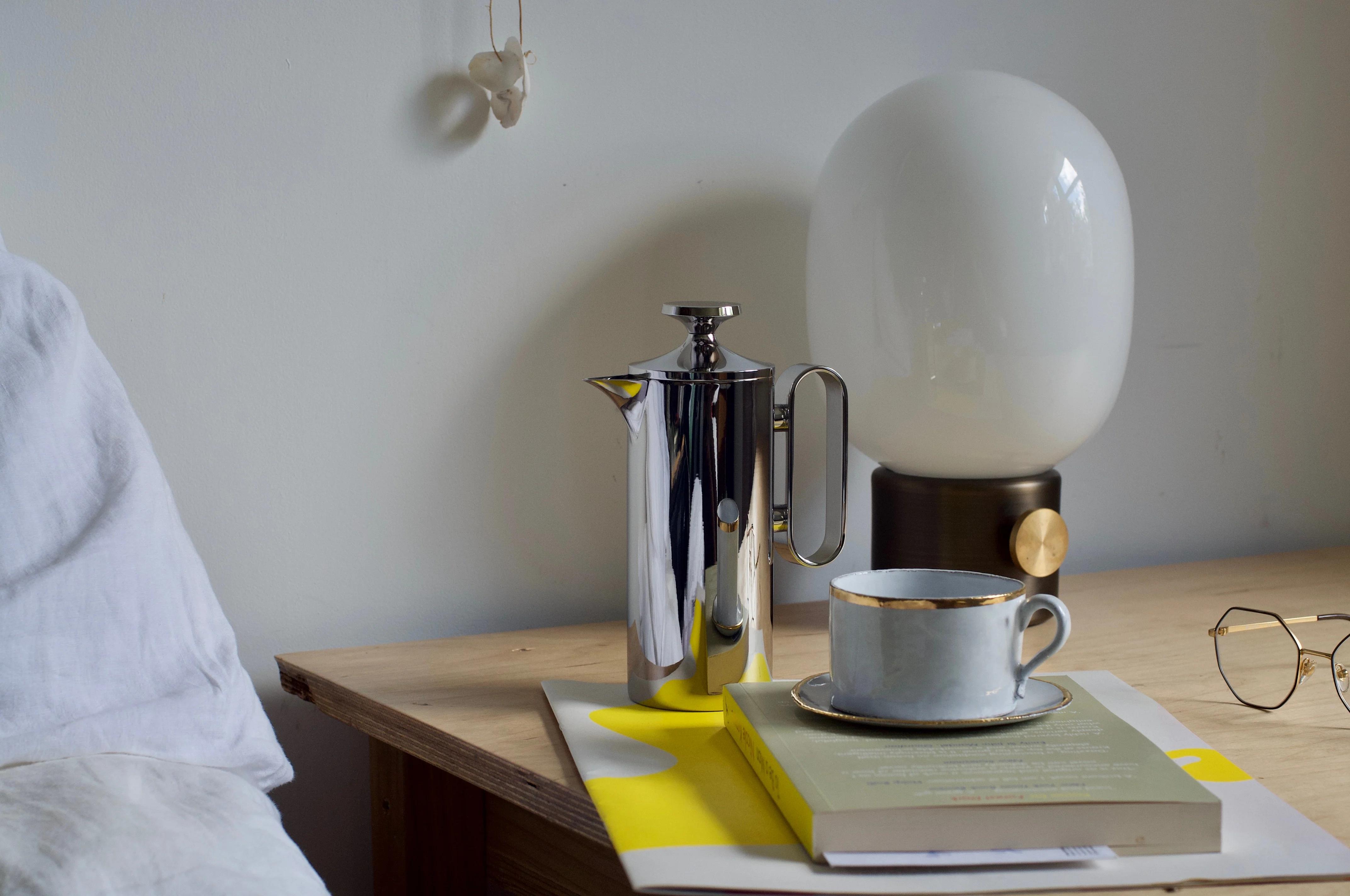 Bedside table with David Mellor cafetière, grey gold-rimmed teacup on saucer atop book and magazine. Modern spherical lamp and glasses in background.