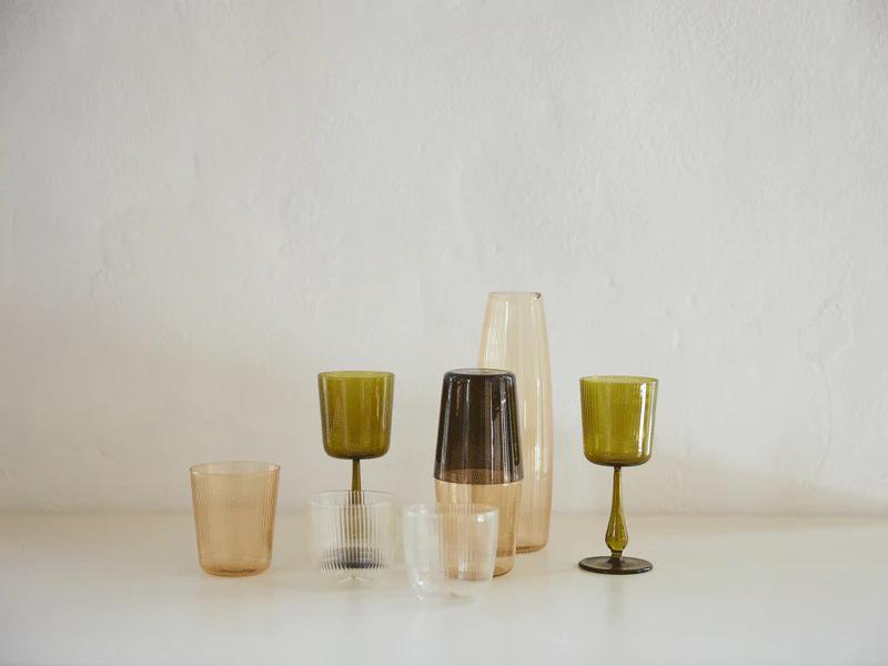 Assorted R+D.LAB glassware on white surface against textured wall. Brown, green, and clear pieces in various sizes showcase studio's research-inspired designs.