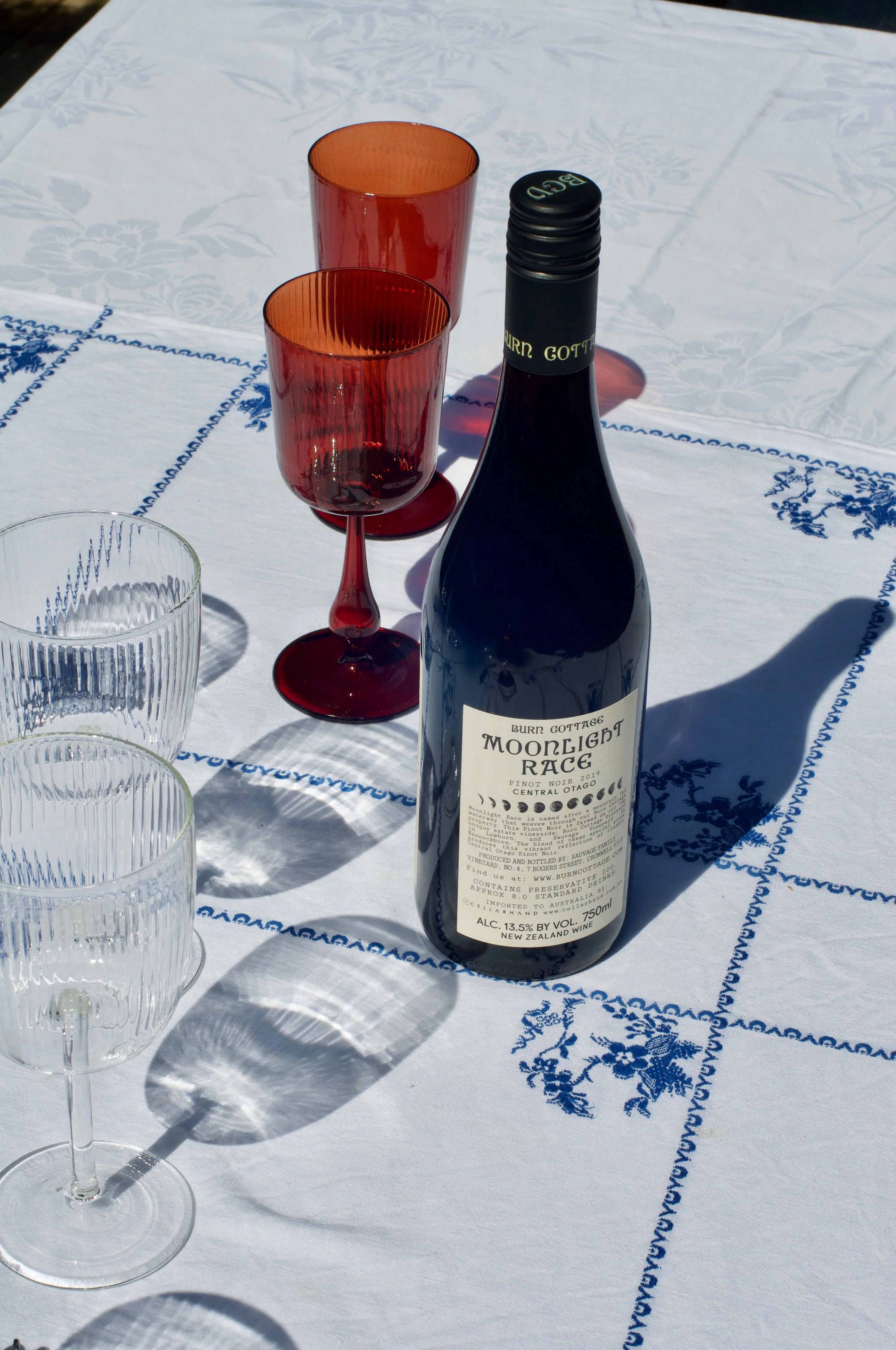 "Moonlight Race" wine bottle on embroidered tablecloth. Two clear and two red-tinted wine glasses surrounding bottle.