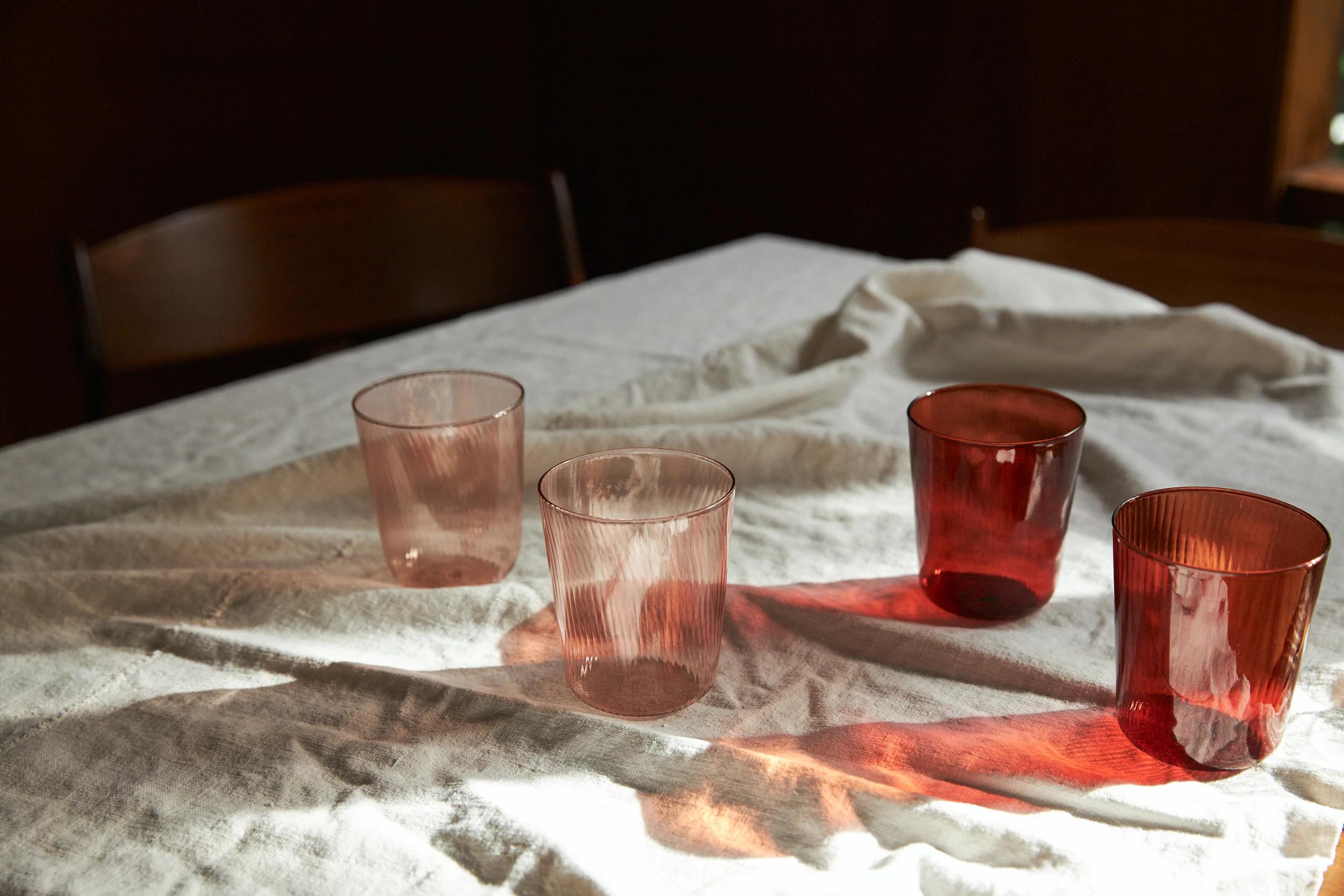 Four coloured glasses on a white wrinkled tablecloth with sunlight casting soft pink and red shadows from the glassware over the fabric.