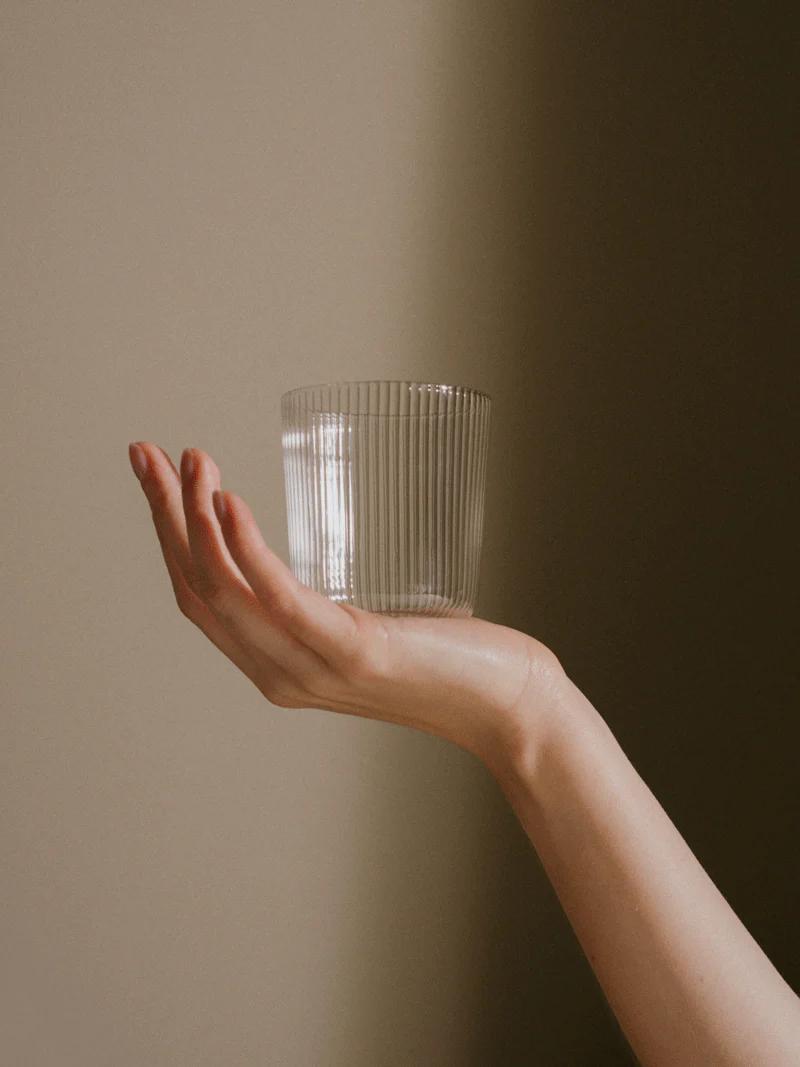 Close-up of ribbed glass cup on outstretched hand against beige background. Horizontal arm creates elegant composition, embodying R+D.LAB's refined style.