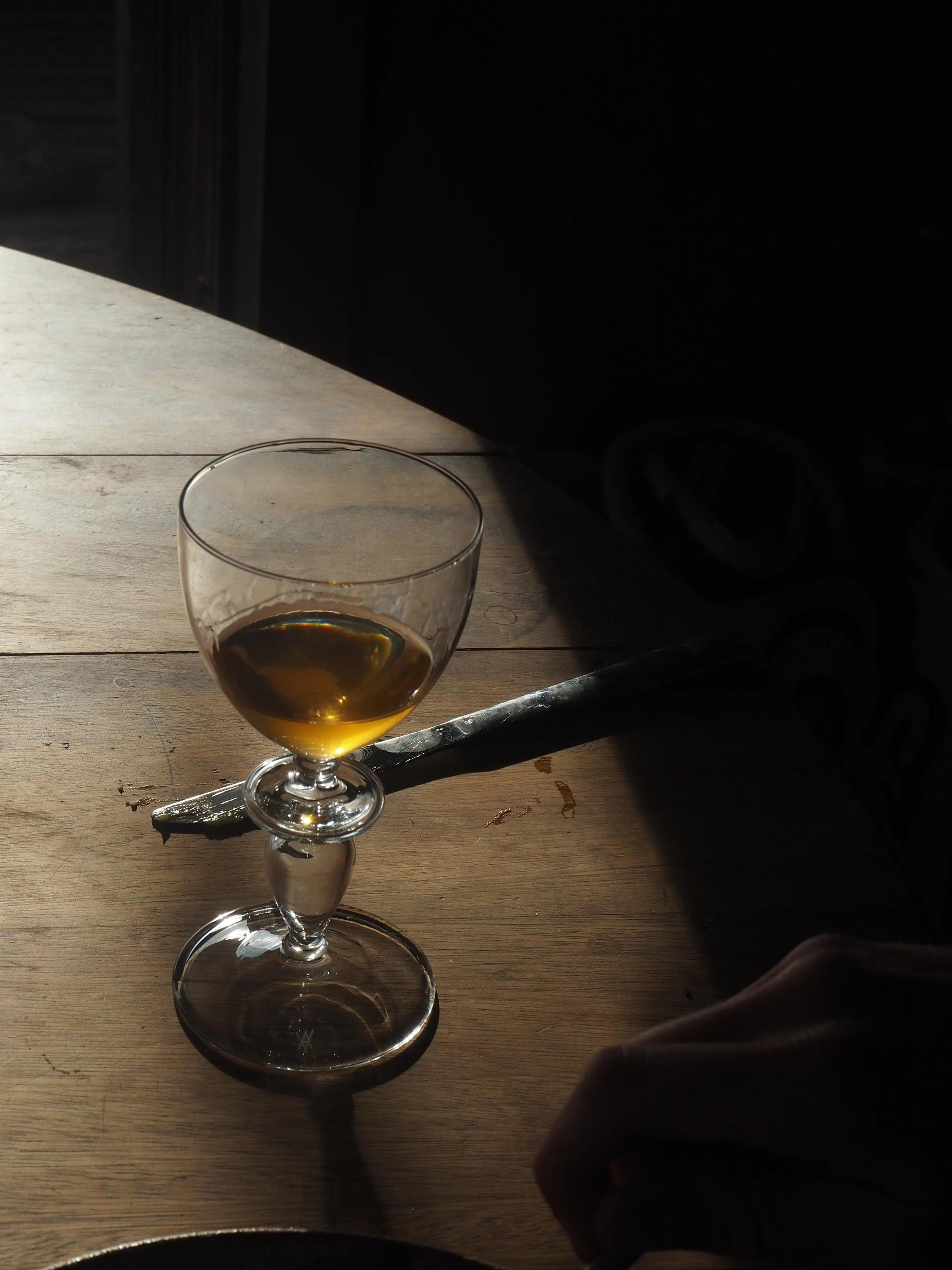 Amber natural wine in glass on wooden table in soft light. Metal object beside glass. Partial view of person's hand in corner. Warm, serene atmosphere.