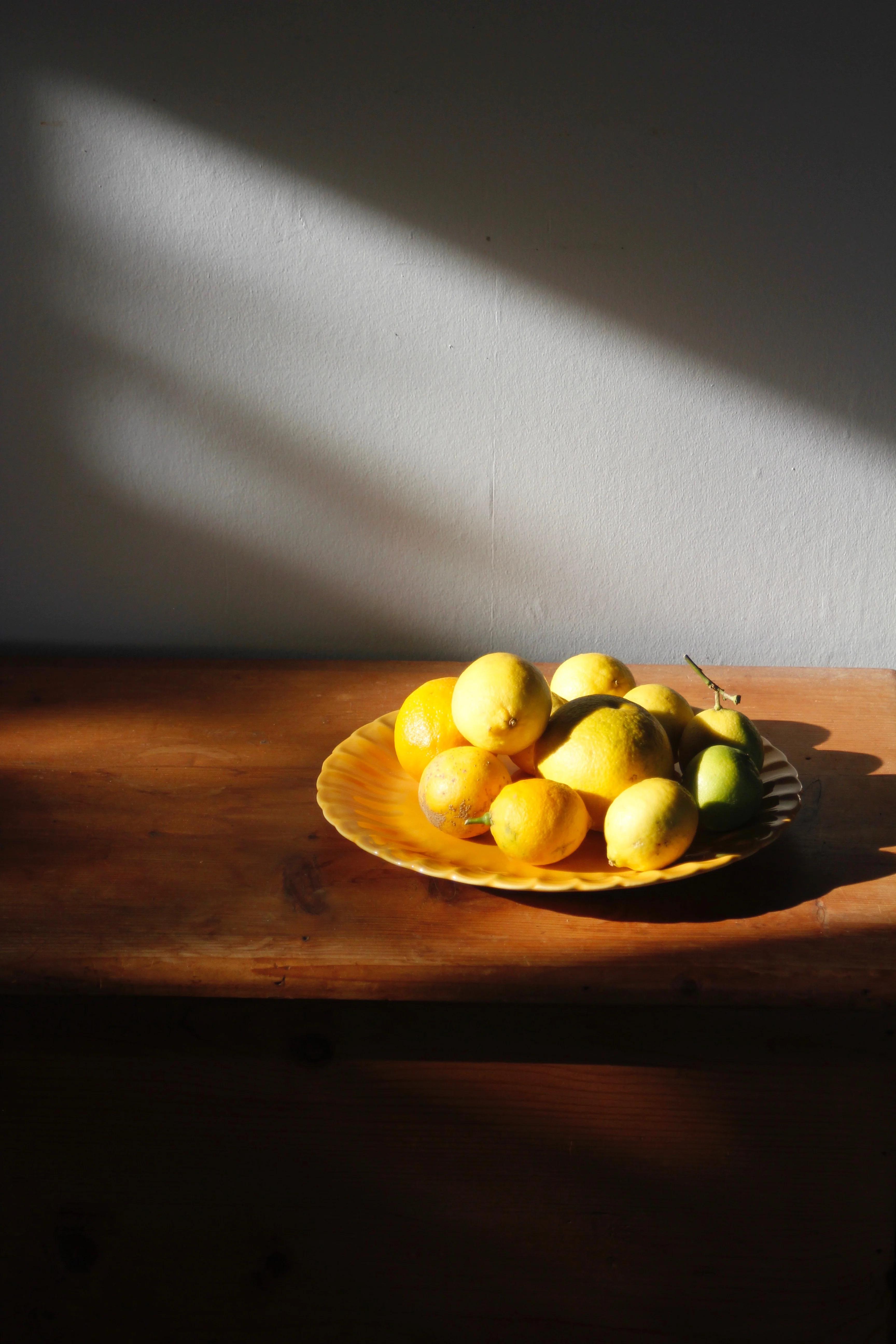 Yellow plate with lemons on wooden surface, surrounded by vibrant tableware. Soft sunlight casts shadows on white wall, creating serene atmosphere.