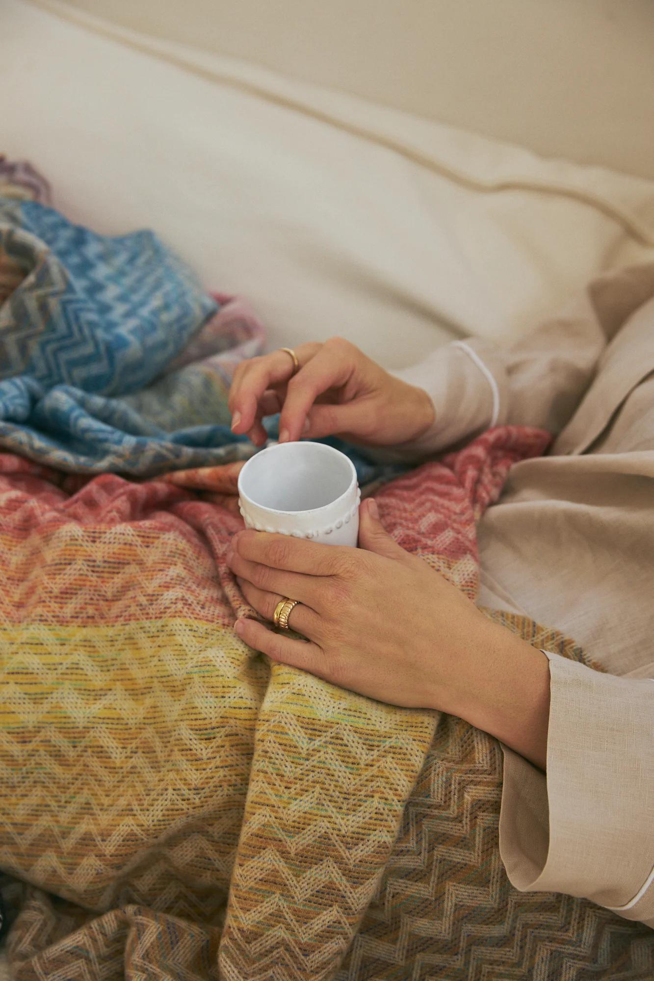 Person holding white mug under zigzag blanket. Wearing beige clothing. Cozy setting.