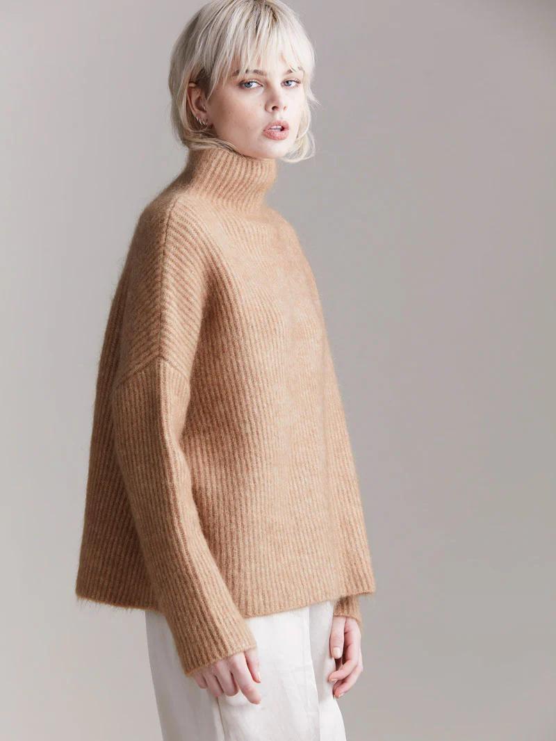 Person with short blonde hair in camel turtleneck sweater. Looking at camera with neutral expression against light background.