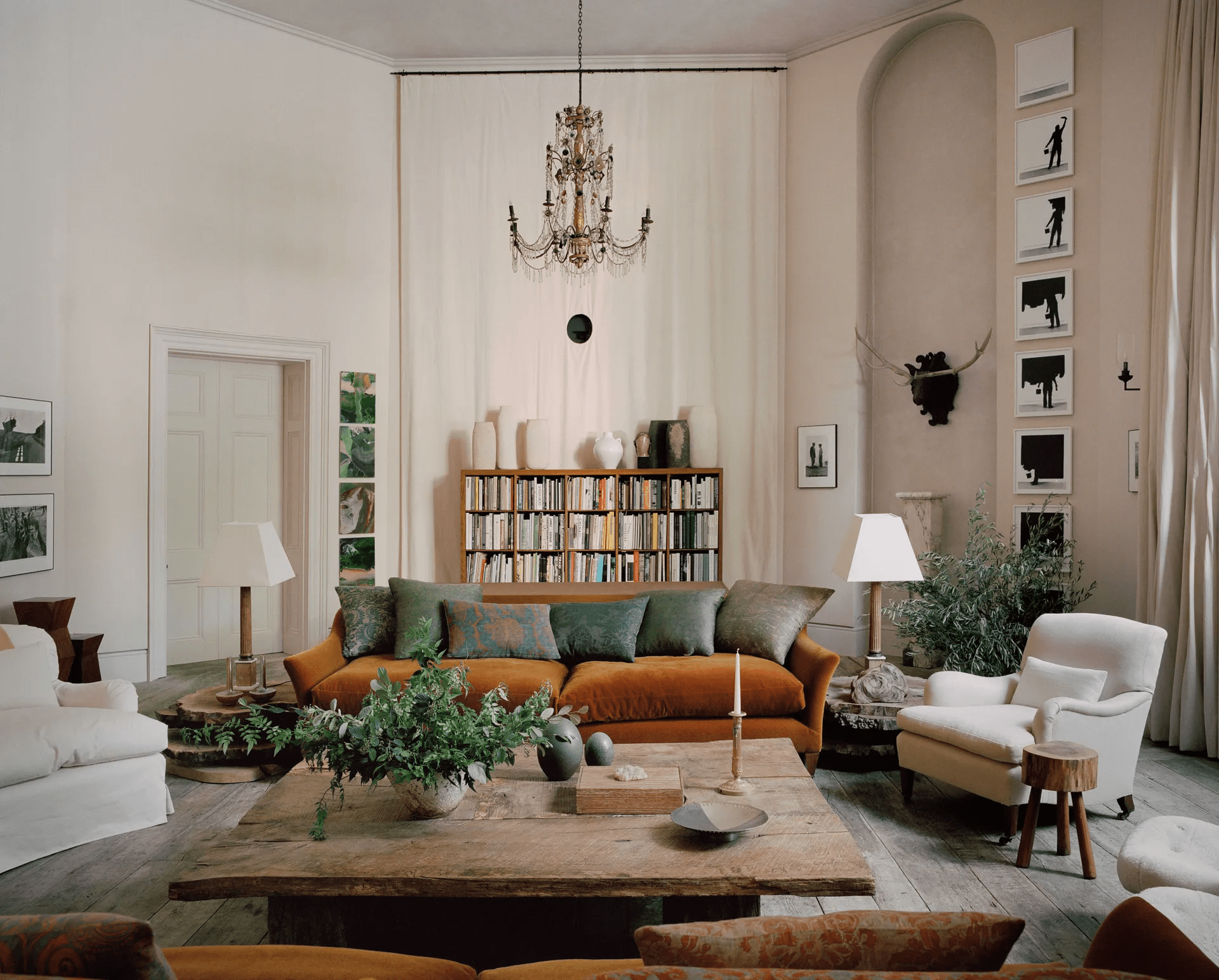 Living room with orange velvet sofa, white armchairs, wooden coffee table. Bookshelf and chandelier.