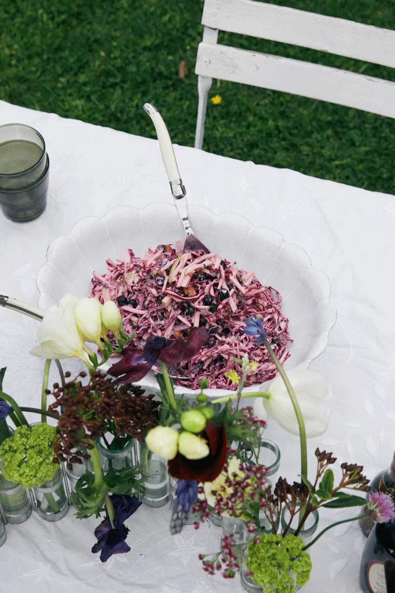 Colourful coleslaw bowl on white tablecloth. Flowers and green glasses on chair, set on lawn.