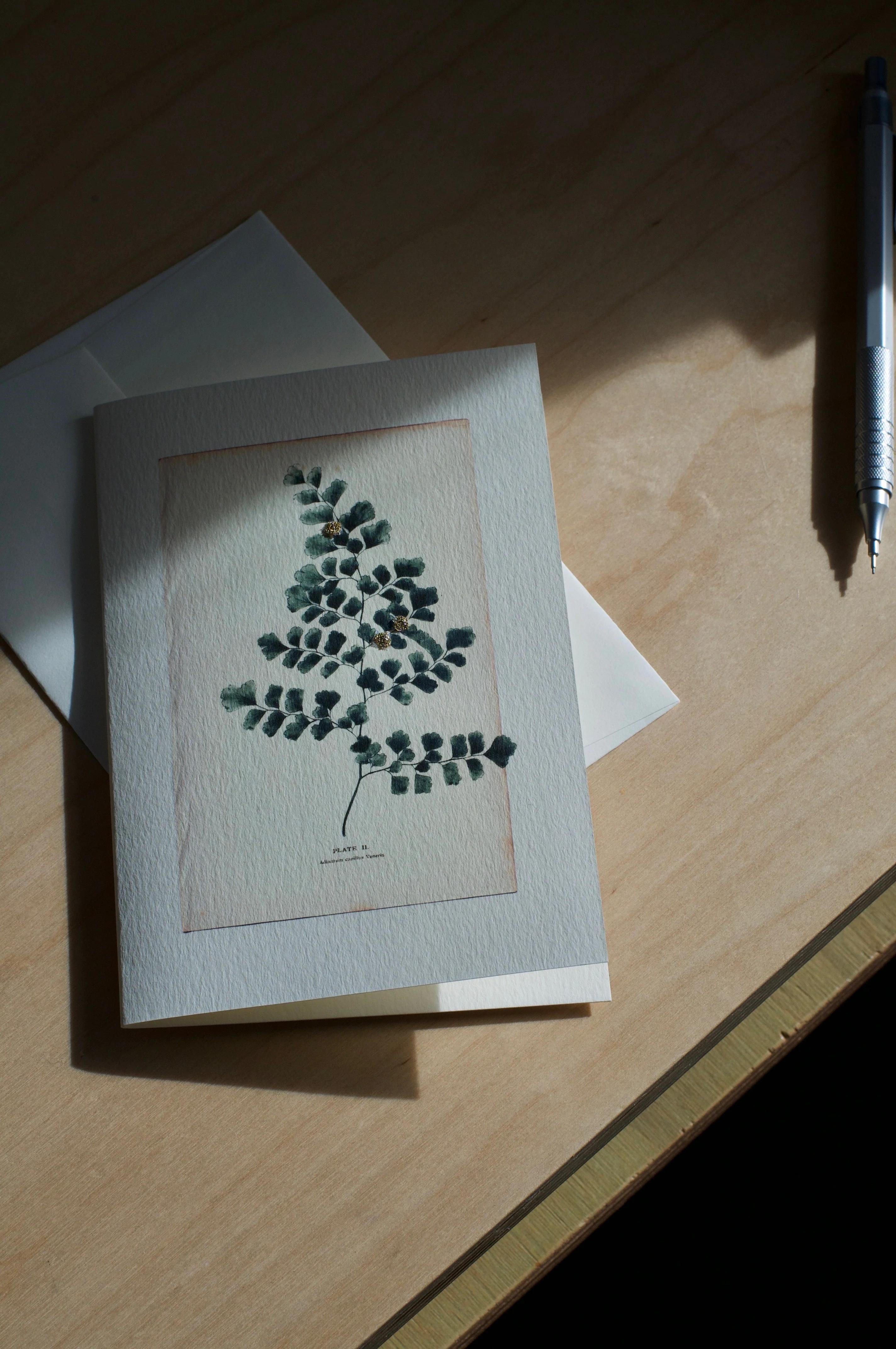 A greeting card featuring pressed leaves and a flower on a wooden surface. A pen lays to the right and sunlight casts a shadow over the scene.