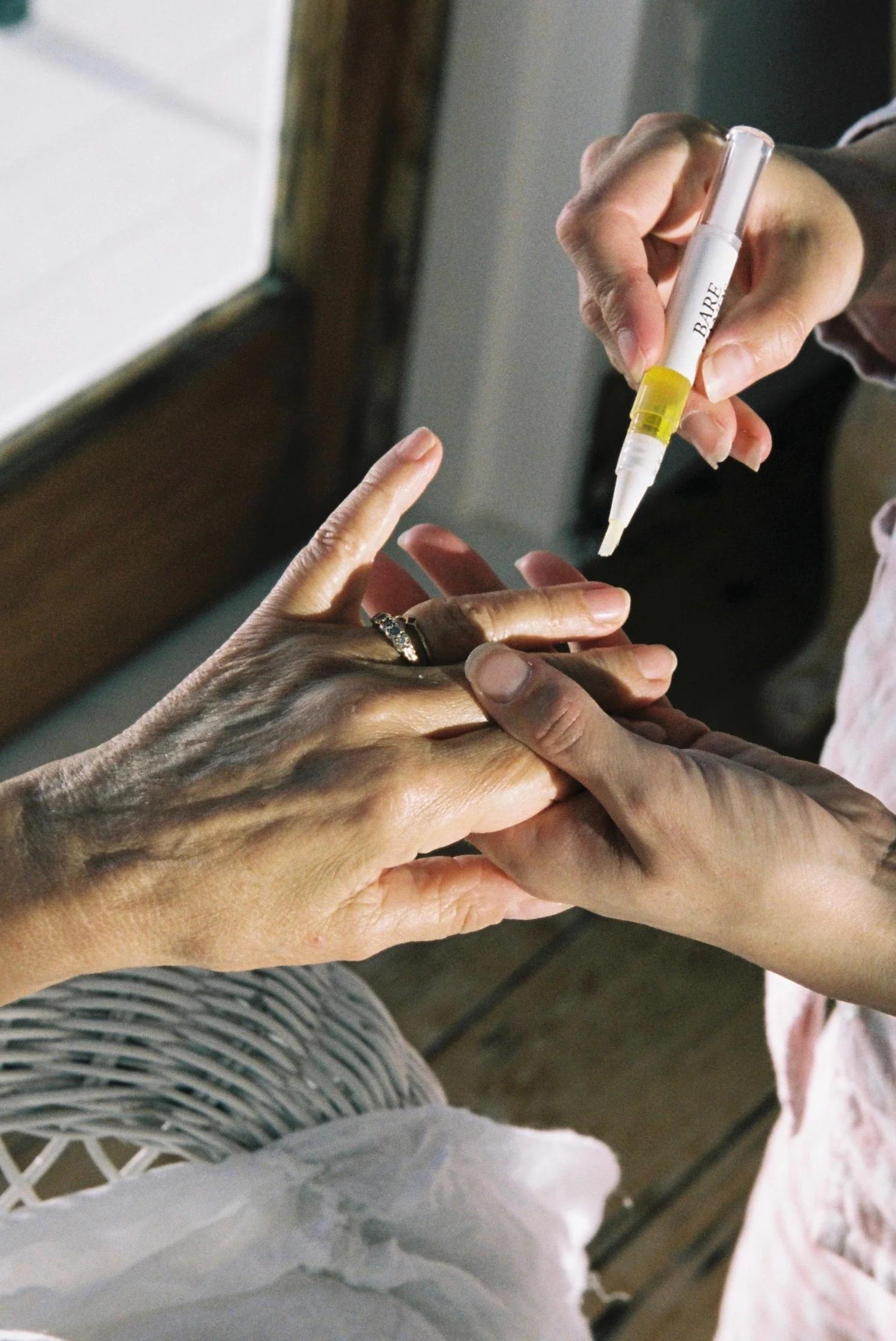 Close-up of two pairs of hands. One person applies yellow cuticle oil to the other’s nails with a pen-like applicator. A window and woven basket are in the background.