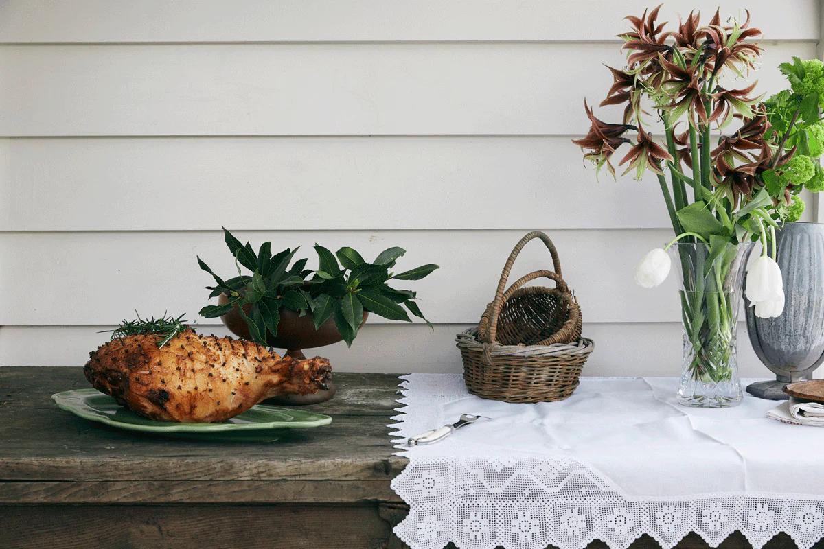 A rustic wooden table with a roast ham, fresh green produce, and a vase of tulips. A white lace tablecloth adds charm.