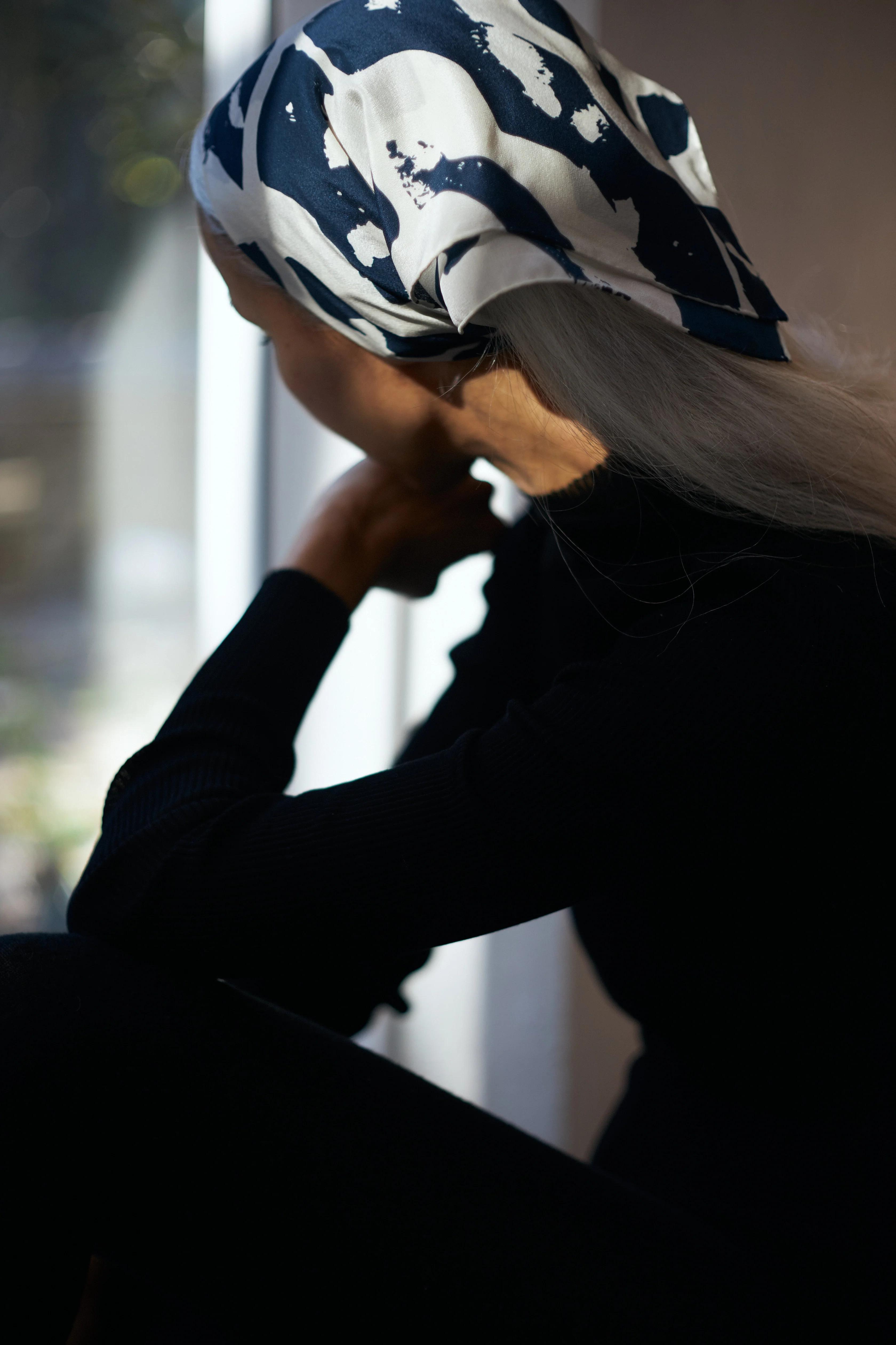 A person with long hair, wearing a black top and a navy and white patterned headscarf, sits pensively by a window with sunlight streaming in. Their head is turned away, chin resting on their hand, embodying thoughtful stillness.