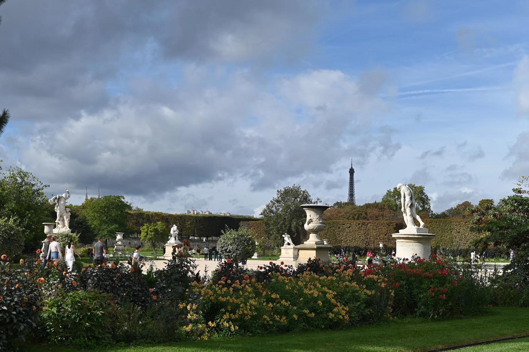 Lush garden with colourful flowers and statues. Eiffel Tower visible above treeline. Visitors strolling through park.