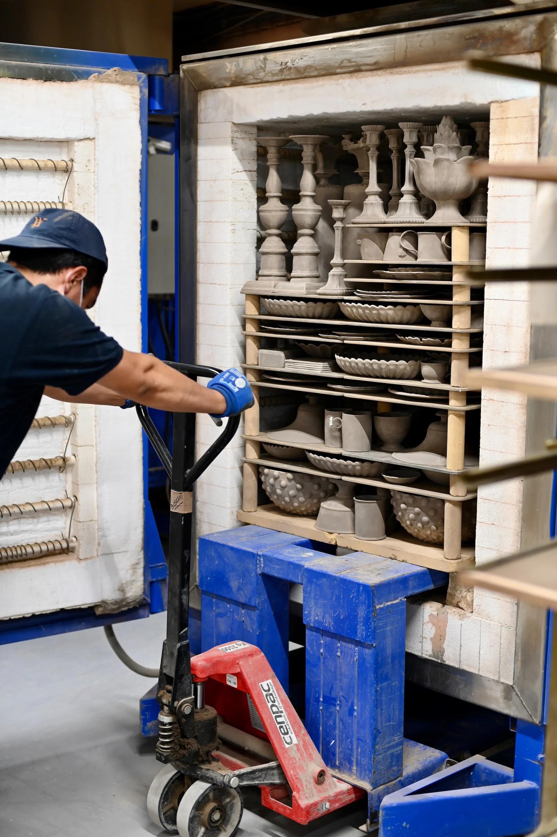 Person operates pallet jack moving loaded kiln with ceramic items. Kiln filled with unglazed pottery pieces. Person wears cap, face mask, gloves, and dark shirt.