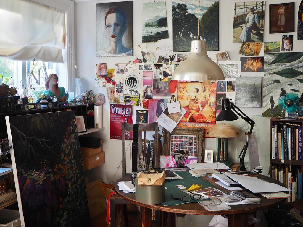 Bustling artist's studio with artworks and posters. Desk under large lamp covered in supplies. Window overlooks Devonport, streaming natural light in.