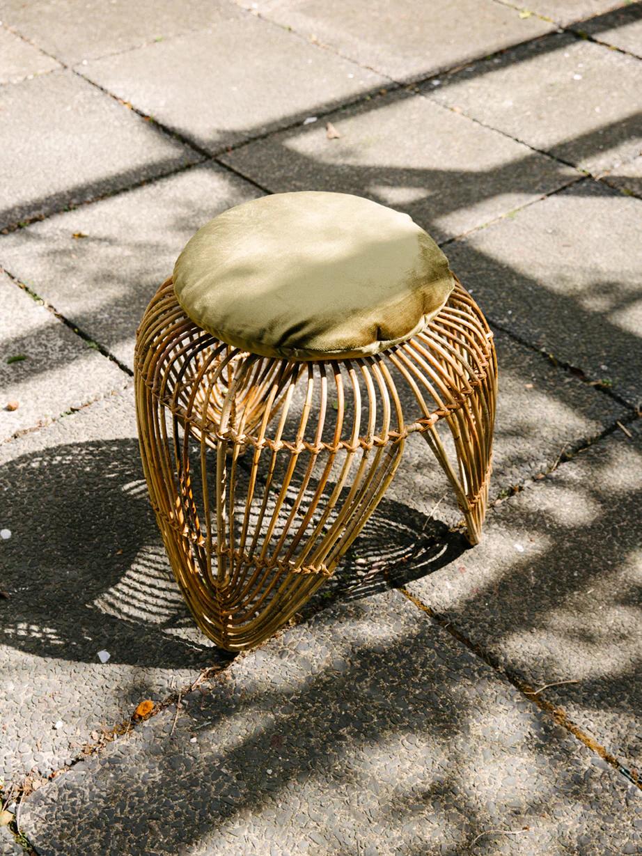 Round wicker stool with green cushion on stone patio. Dappled shadows create checkered pattern. Open design suggests lightweight, portable KLAY piece.