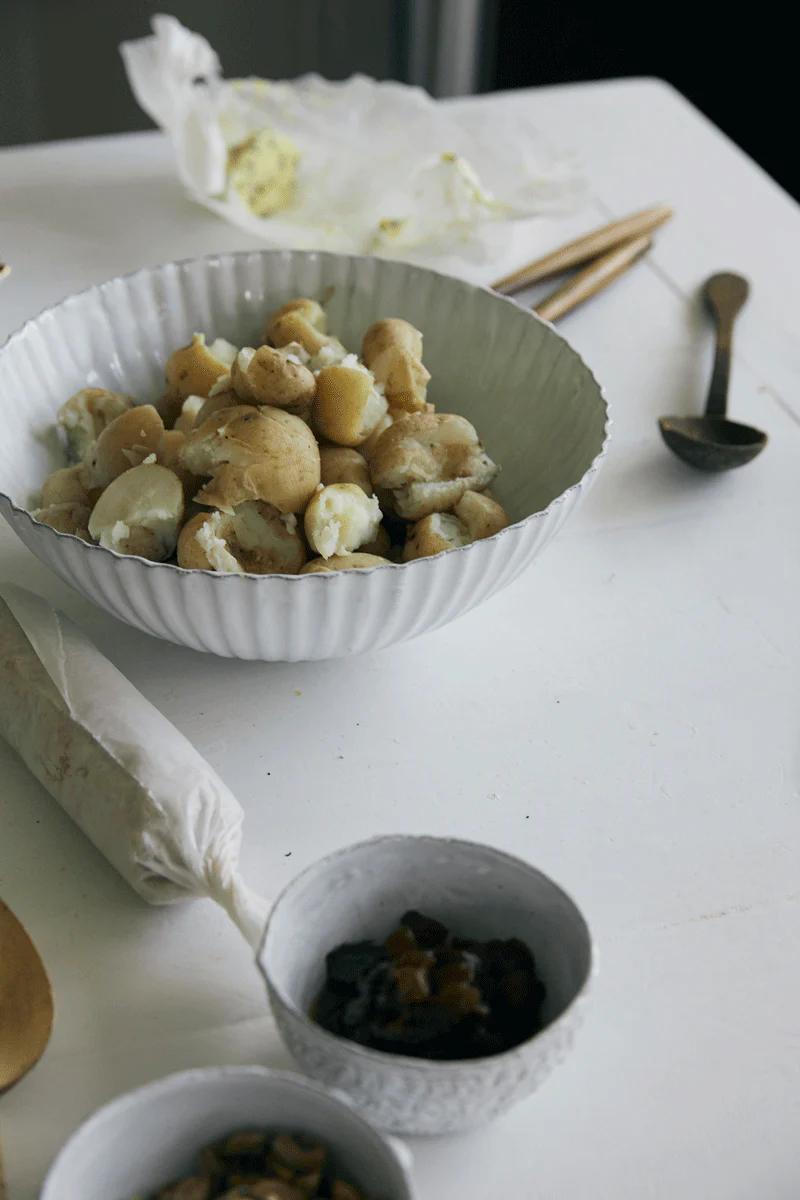 A white bowl of boiled baby potatoes on a white table. Nearby are nuts, dried fruit, butter, cheese, and chopsticks—perfect for summertime gatherings with Tessuti family recipes.