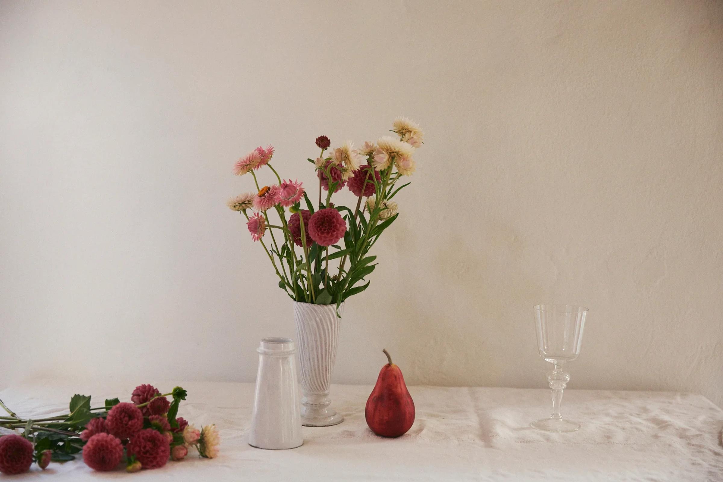 White vase with pink and cream flowers, red pear, empty wine glass, and small container on white tablecloth. Scattered flowers to left.