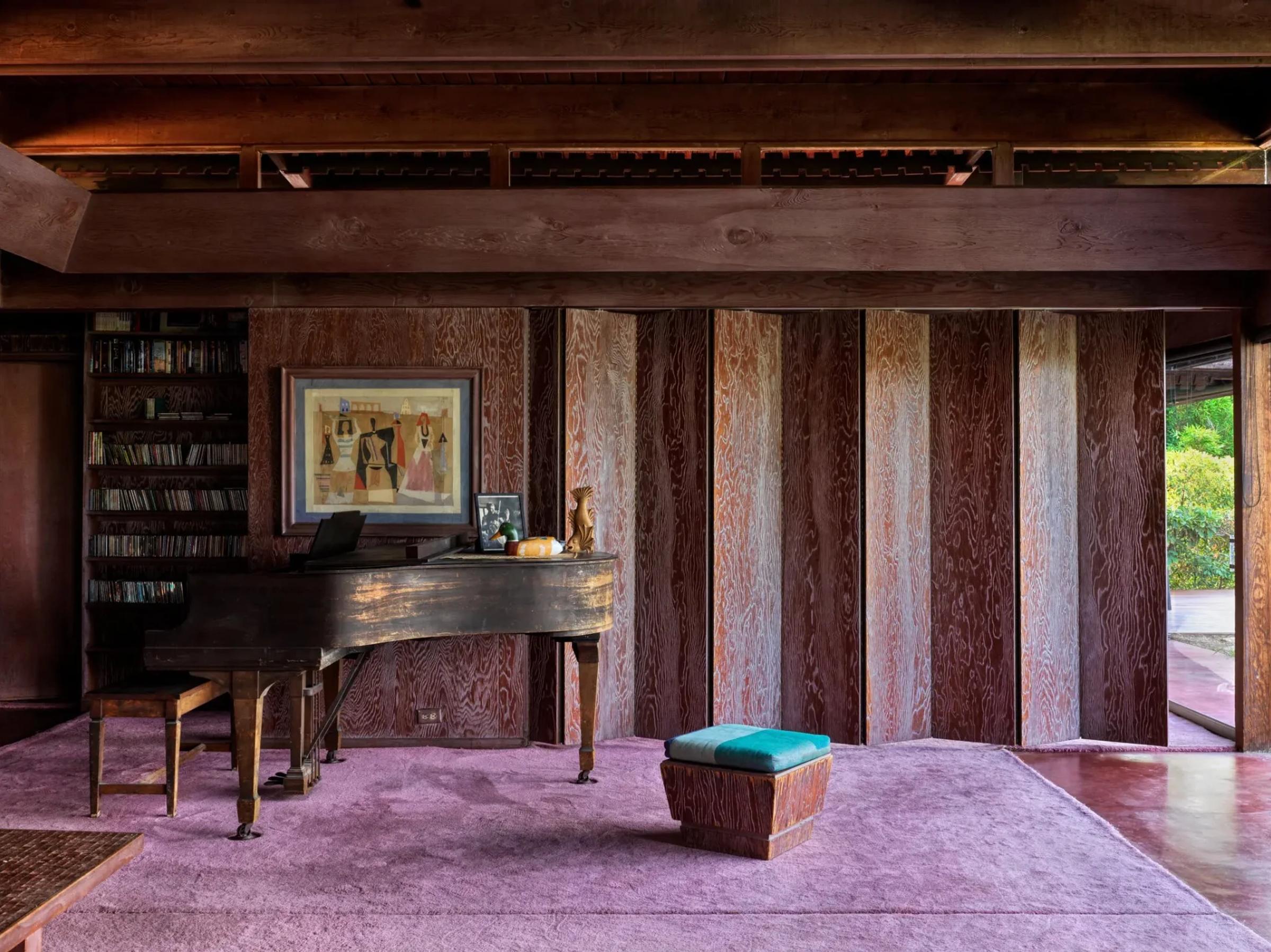 A spacious room with angular wooden panel walls, purple carpet and a large window. Features a vintage grand piano with art and decor on top, small teal stool, and book shelf in back left corner.
