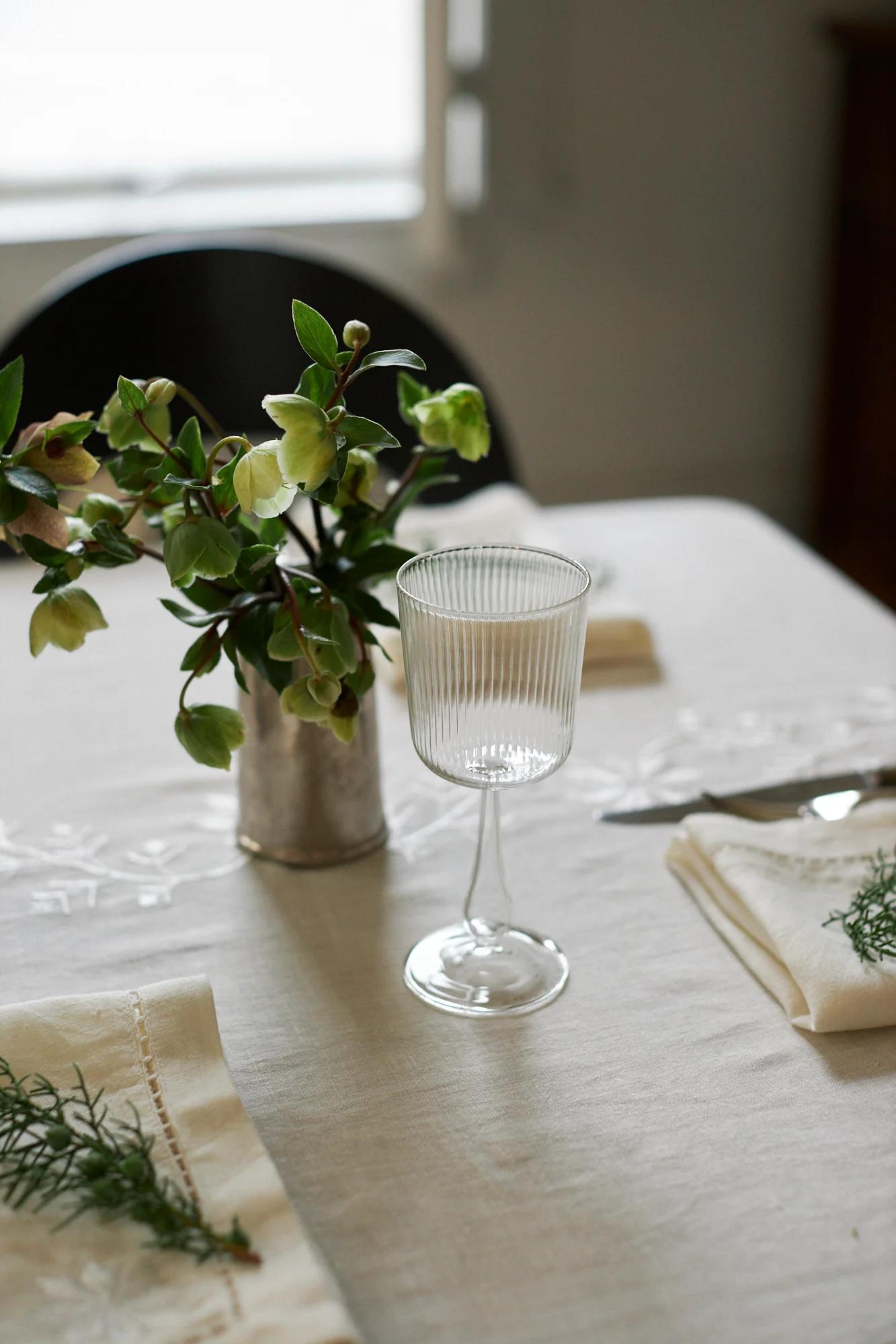 A table set with a white cloth, ribbed glasses, and napkins with greenery. A small vase of flowers serves as a festive centrepiece.