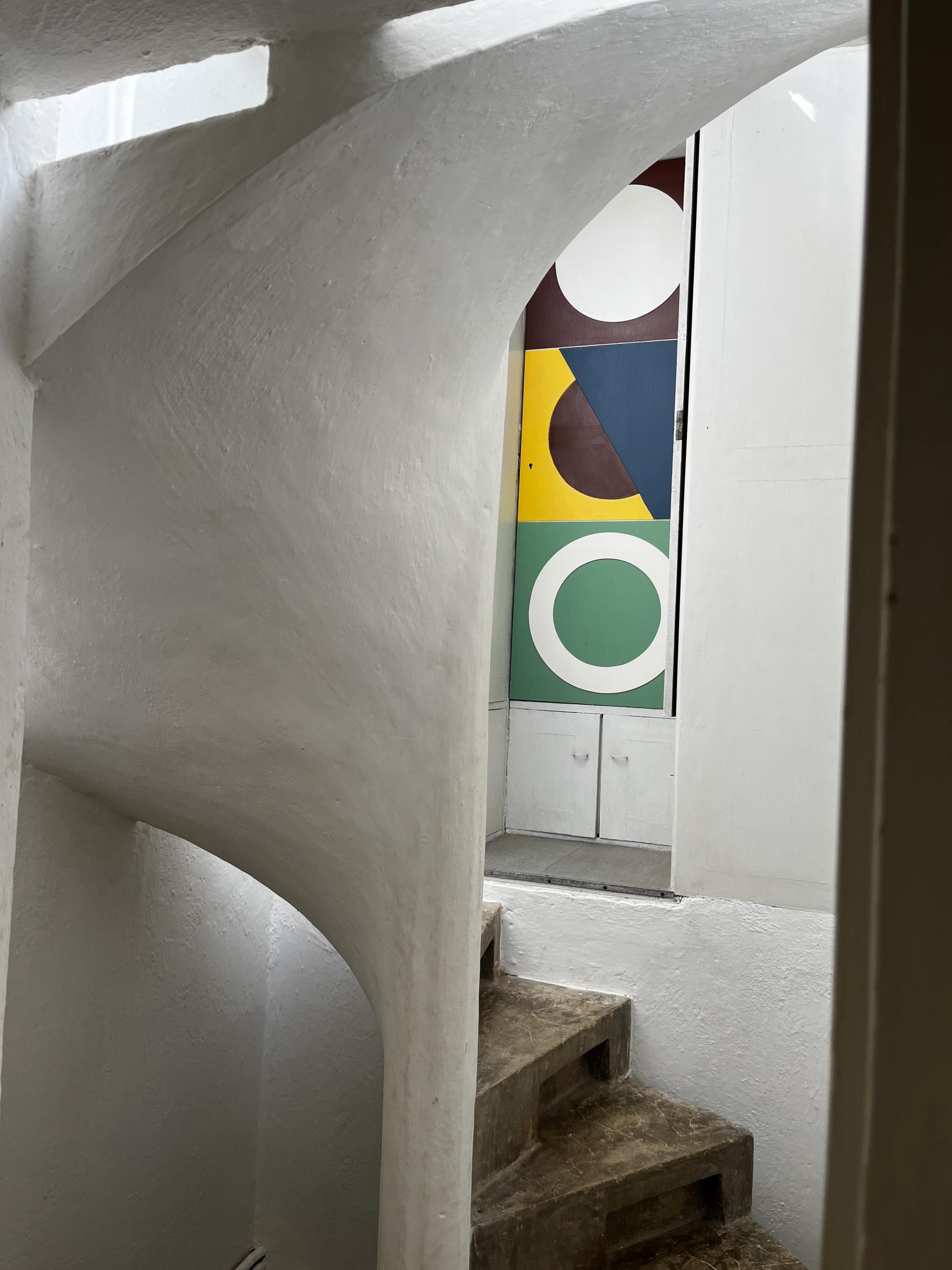 Concrete spiral staircase leads to door with colourful geometric design. White-walled interior.