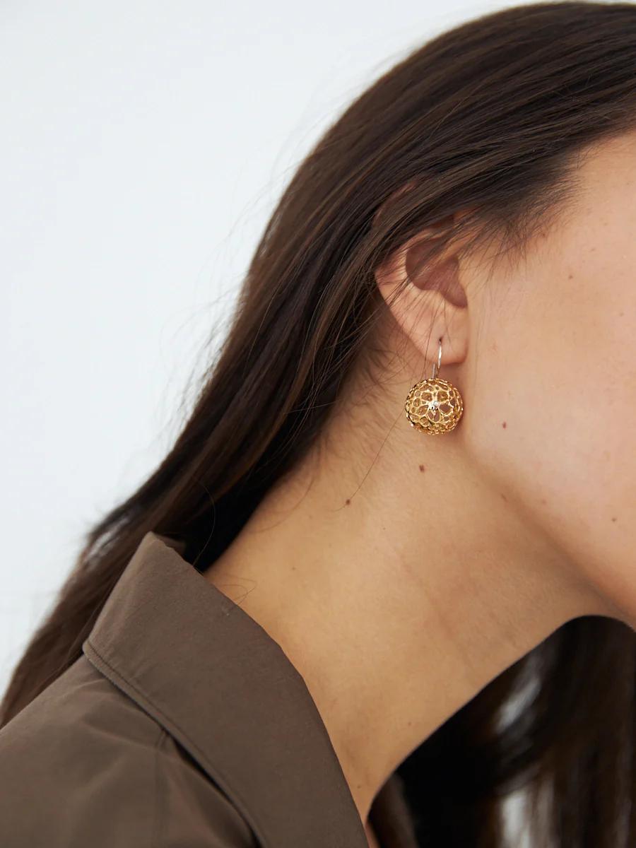 Close-up of woman wearing gold dangling earring. Long, dark hair behind ear. Brown shirt visible.