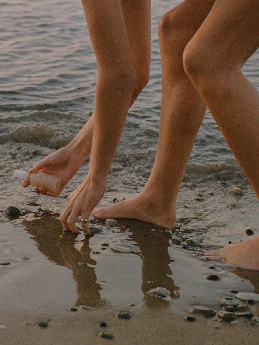 Two people at water's edge on rocky beach. One crouched, holding bottle and touching wet sand.