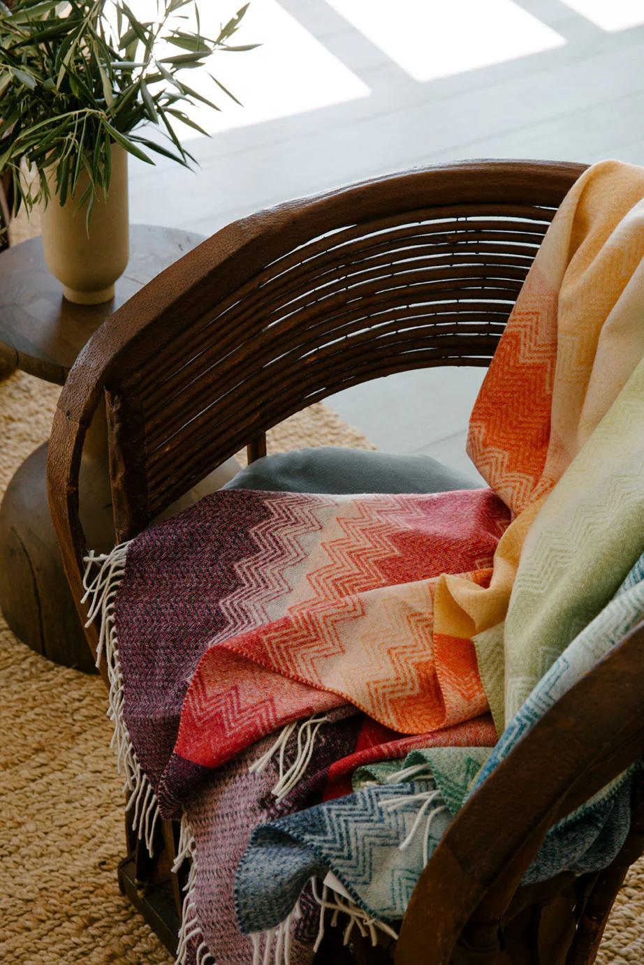 Wooden chair with colourful Missoni-style zigzag blanket. Small table with plant nearby. Natural light creates cosy atmosphere.