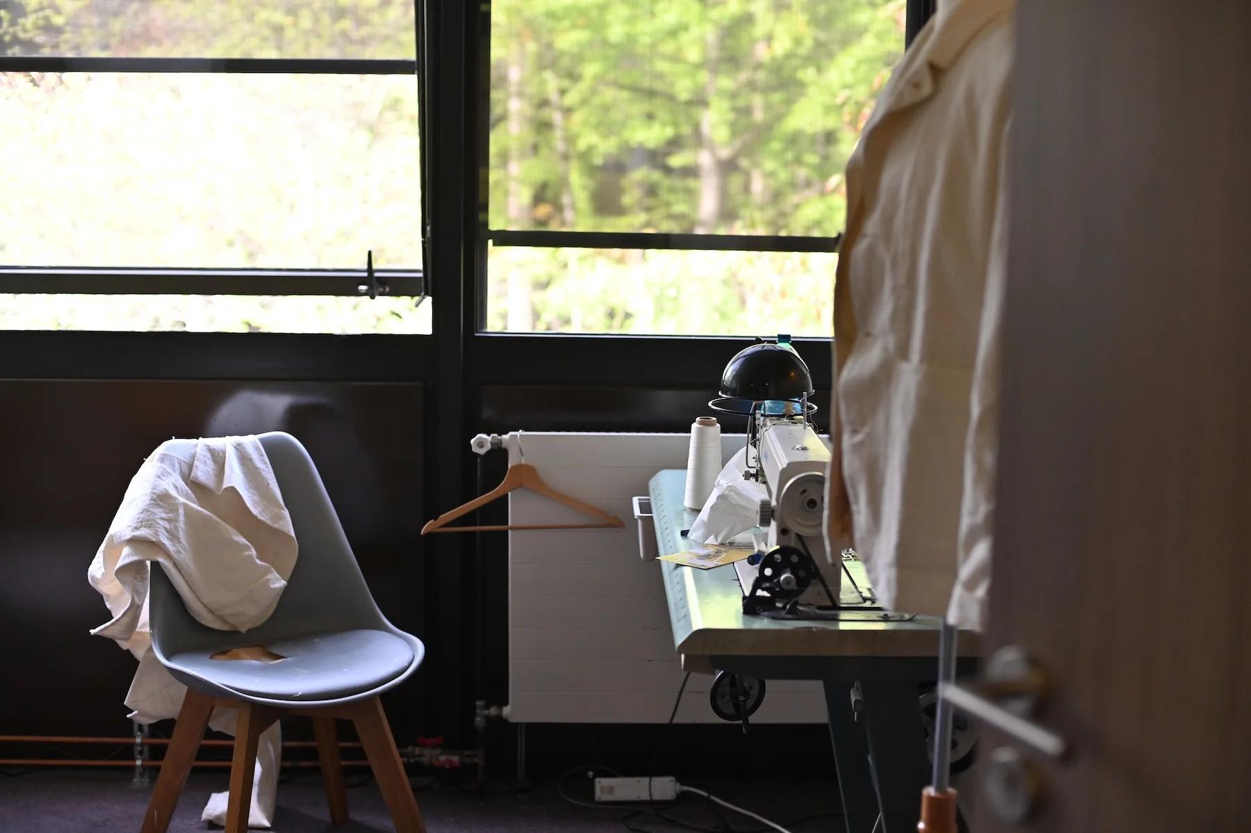 Room with sewing machine, lamp, and fabric-draped chair. Large windows let in natural light.