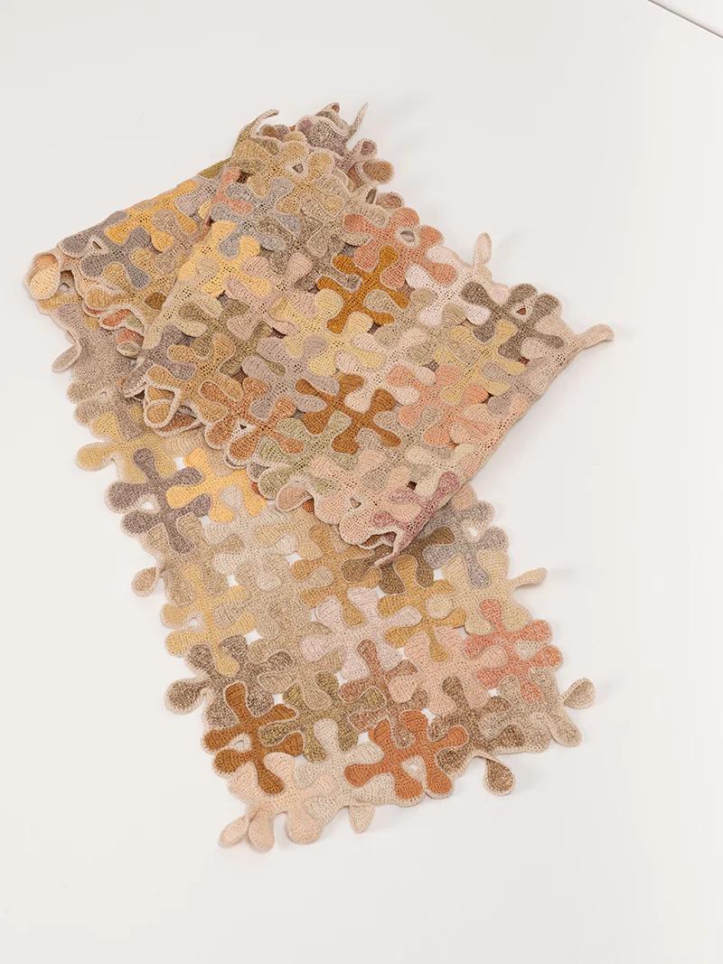 A folded rug with warm hues of beige, brown, pink, and orange, featuring an abstract hand-crocheted pattern by Sophie Digard, placed on a white surface.