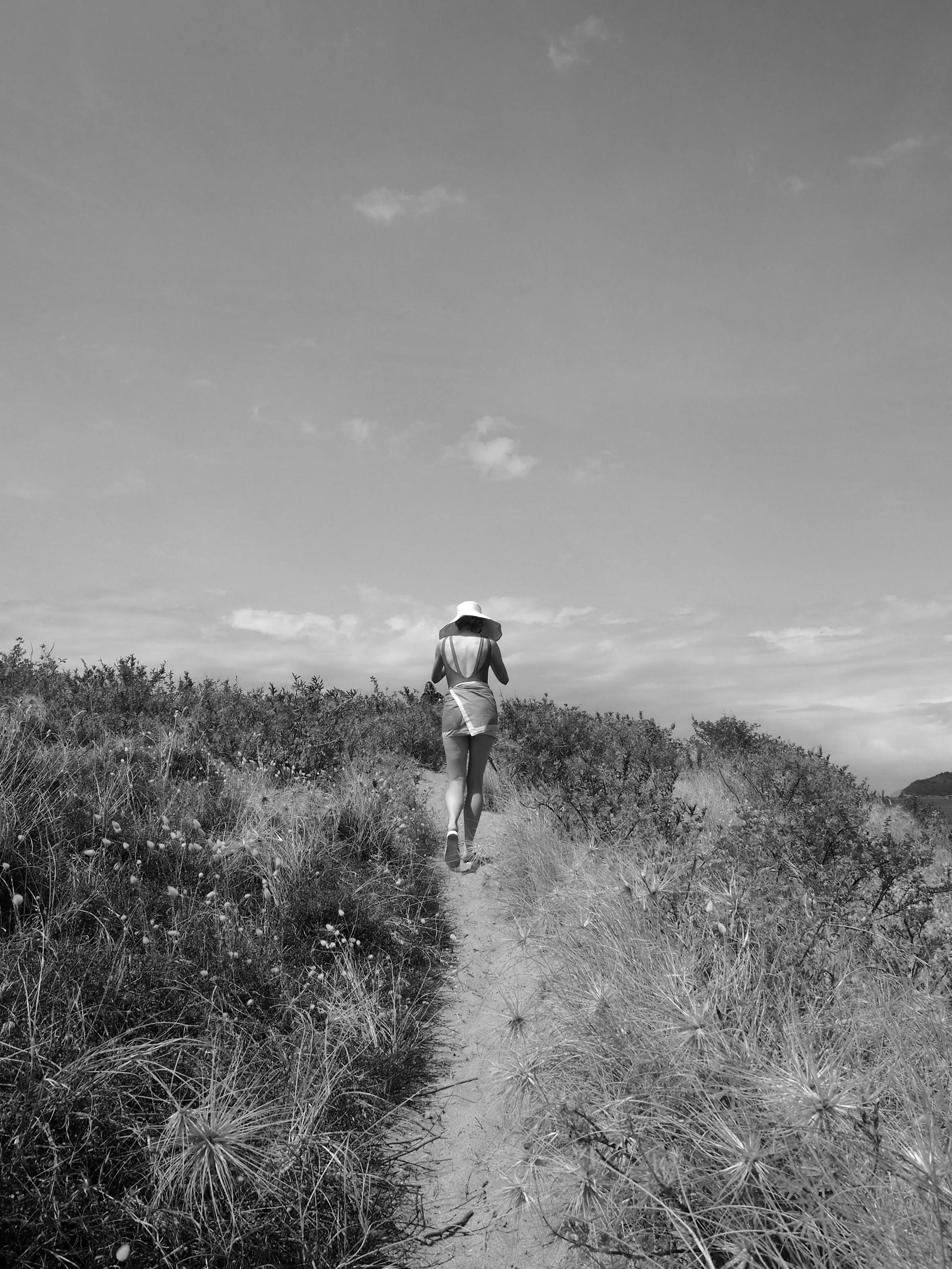 A person wearing a sunhat walks along a narrow path through tall grass toward the beach. The black-and-white scene captures a nostalgic summer moment.