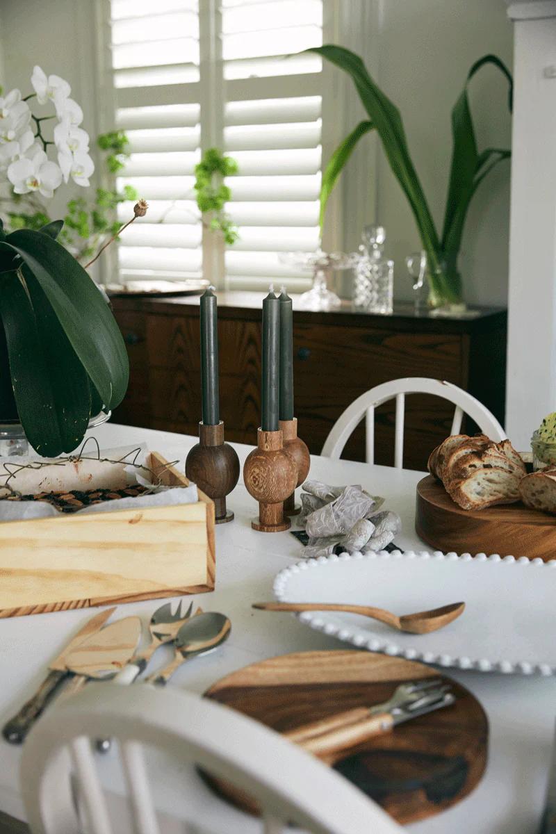 A dining table set with wooden and ceramic items, including bread on a wooden board, cutlery, and two black candles. Green plants and a window with shutters evoke the warmth of the holiday season.