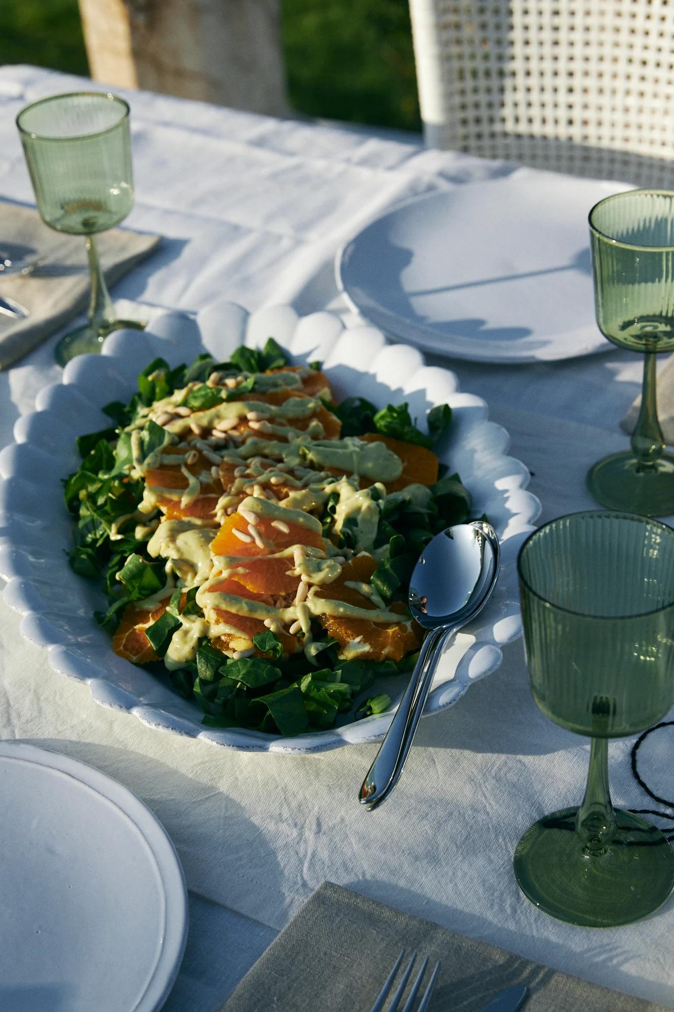 White bowl with salad on outdoor table. Table set with plates, green glasses, cutlery on white tablecloth.